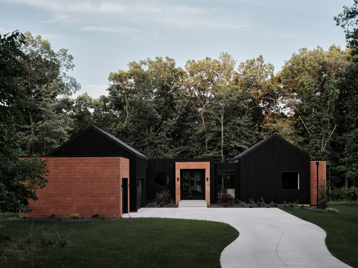 Modern house exterior with black wood siding, terracotta brick walls, a curved concrete driveway, and dense trees in the background.