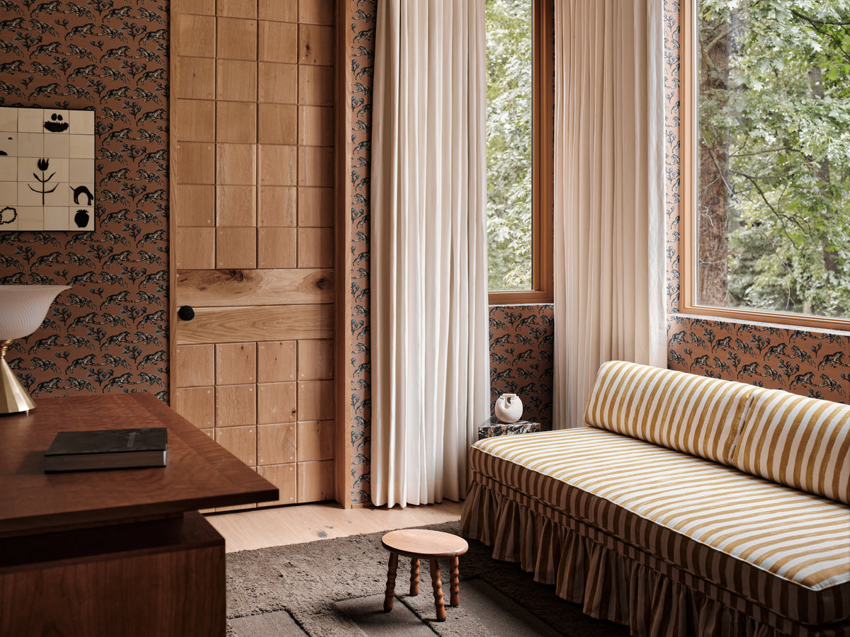 A cozy interior corner with a striped daybed cushion, ruffled skirt, tall windows, cream curtains, patterned wallpaper, and a wooden door. A small stool sits on the floor beside the window.