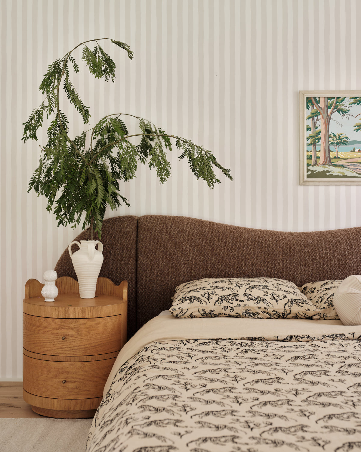 A styled bedroom scene featuring a bed with a brown upholstered headboard, patterned beige bedding, matching pillows, a light wood nightstand, and a tall green potted plant against a cream striped wall.