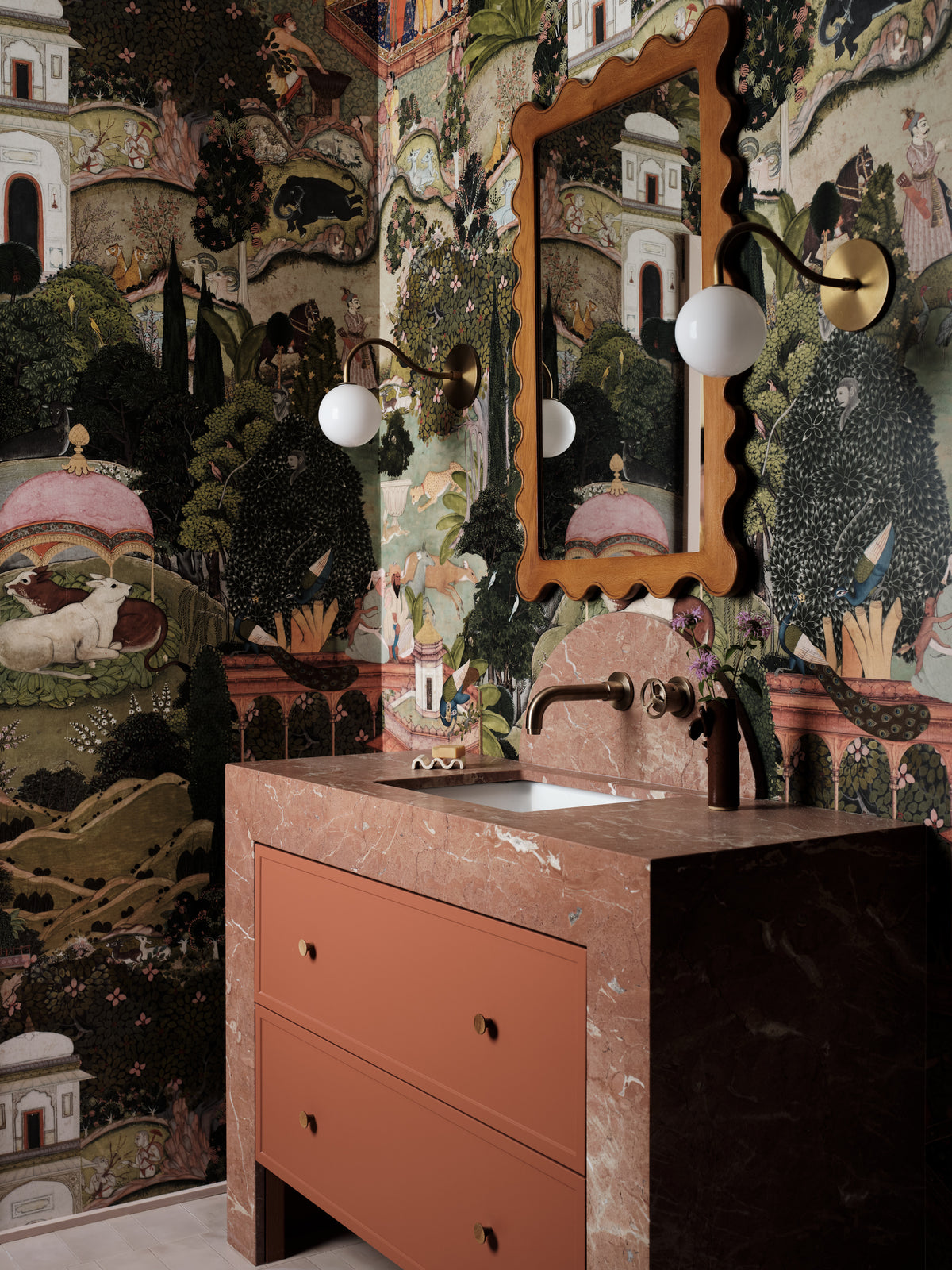 A bathroom vanity setup featuring a terracotta-colored drawer cabinet with a matching stone-look countertop, set against a highly detailed, multicolored landscape wallpaper and an ornate scalloped wooden mirror.