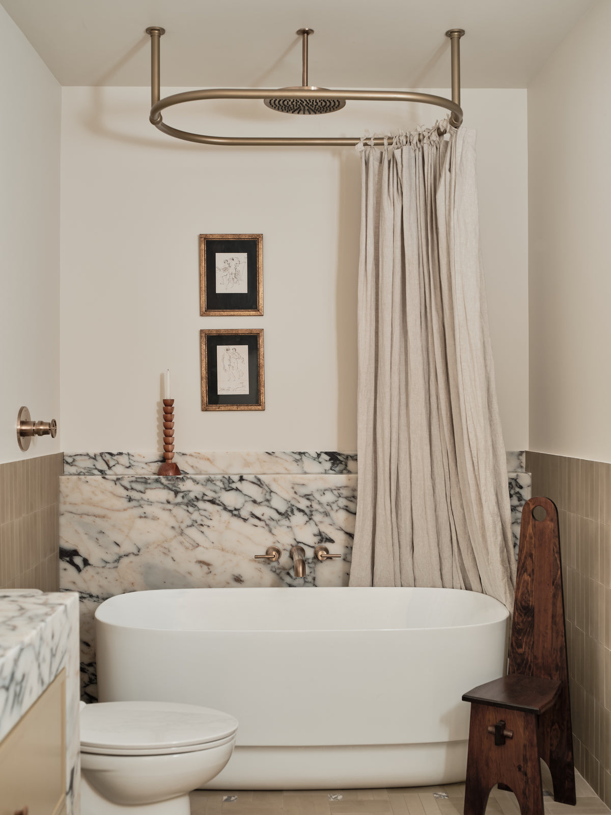 A bathroom interior featuring a white freestanding bathtub, a rustic bronze oval shower fixture with a beige curtain, a marble-tiled backsplash, and a dark wood stool.