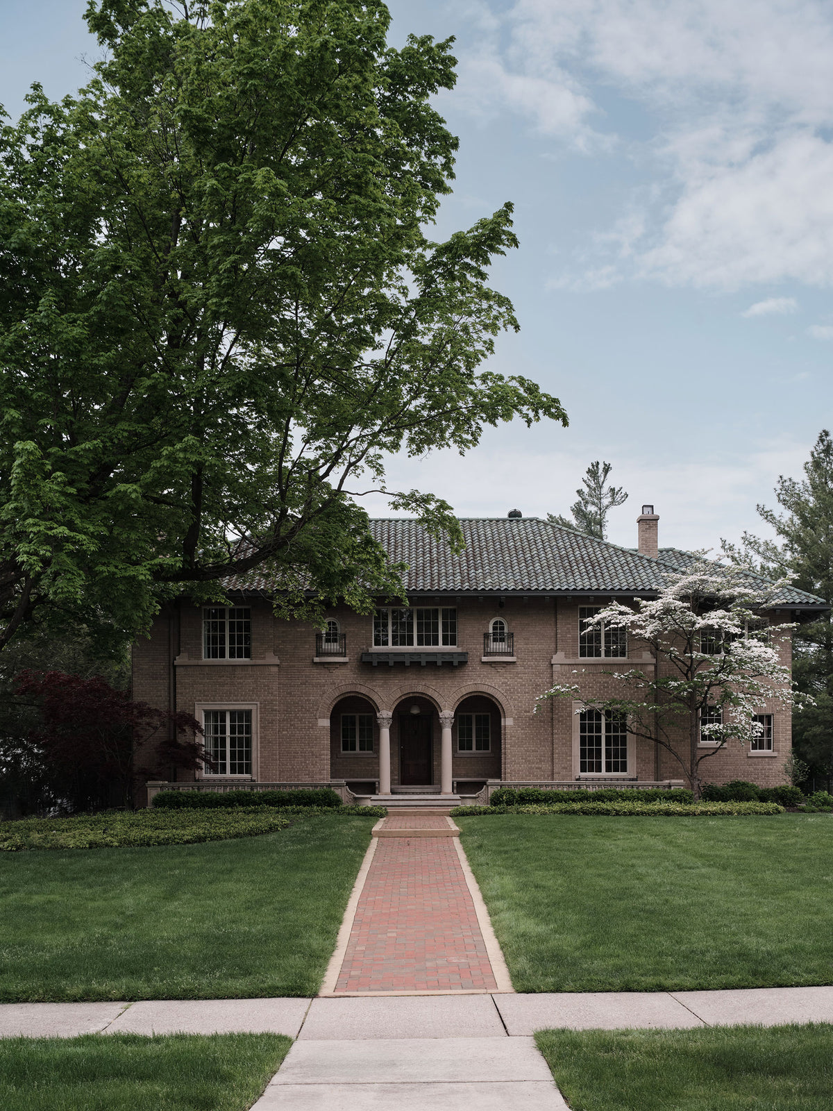 A large brick home with a tiled roof, fronted by a brick pathway leading through a green lawn toward the entrance, with a large tree on the left and a flowering tree on the right under a partly cloudy sky.