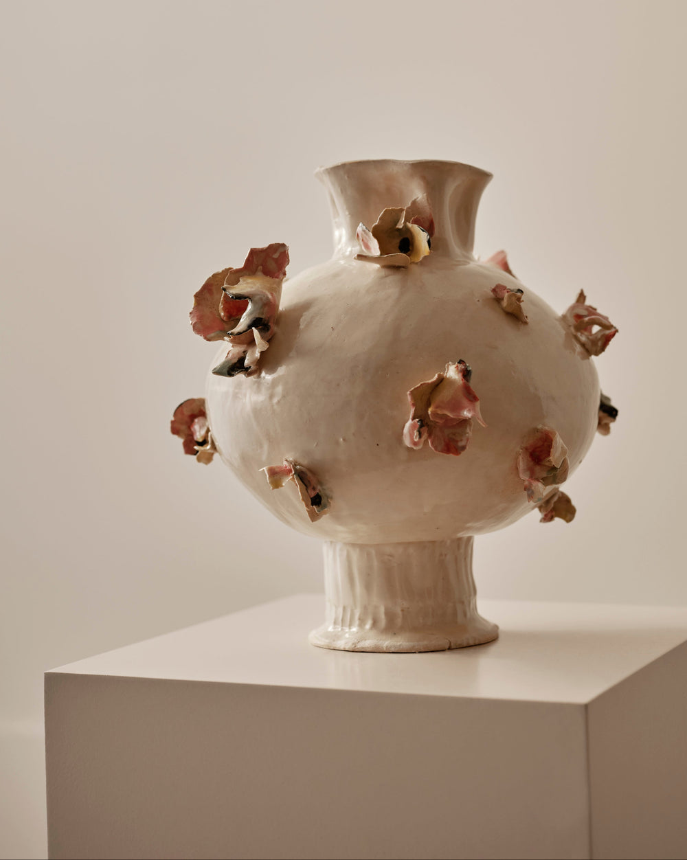 A sculptural ceramic vase with a rounded bulbous body, flared neck, and ribbed pedestal base, shown on a white display pedestal against a warm beige wall. The vase is off-white/cream with multiple small raised floral or petal-like accents in muted pink, terracotta, and olive tones around the body.