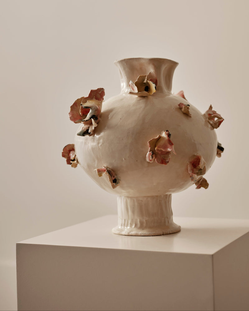 A sculptural ceramic vase with a rounded bulbous body, flared neck, and ribbed pedestal base, shown on a white display pedestal against a warm beige wall. The vase is off-white/cream with multiple small raised floral or petal-like accents in muted pink, terracotta, and olive tones around the body.