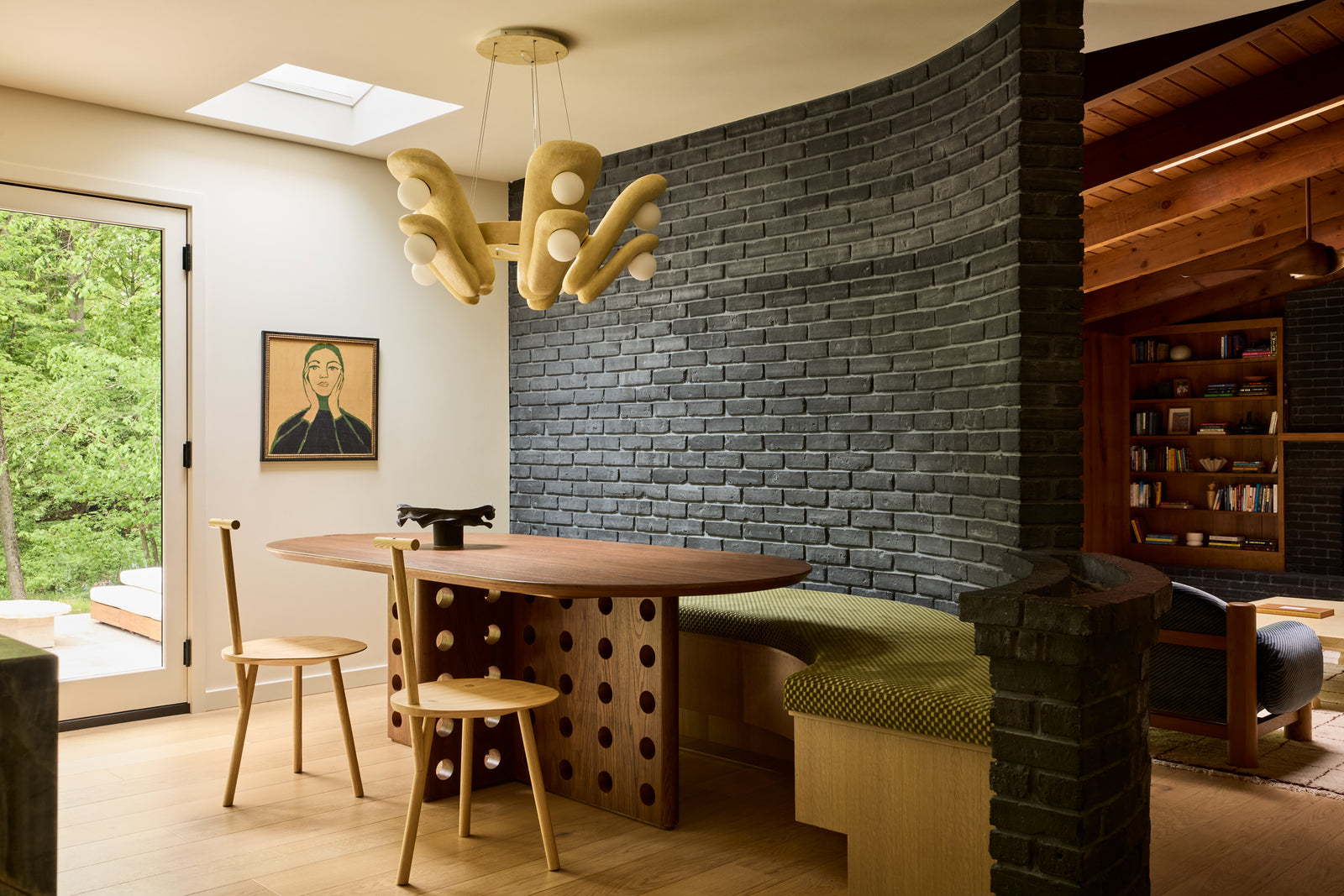 A modern dining nook with a dark gray brick accent wall, a wooden table with a distinctive cutout base, two wooden chairs, and a built-in green bench. A sculptural multi-light pendant hangs from the ceiling, and a large window on the left looks out to a lush green garden.
