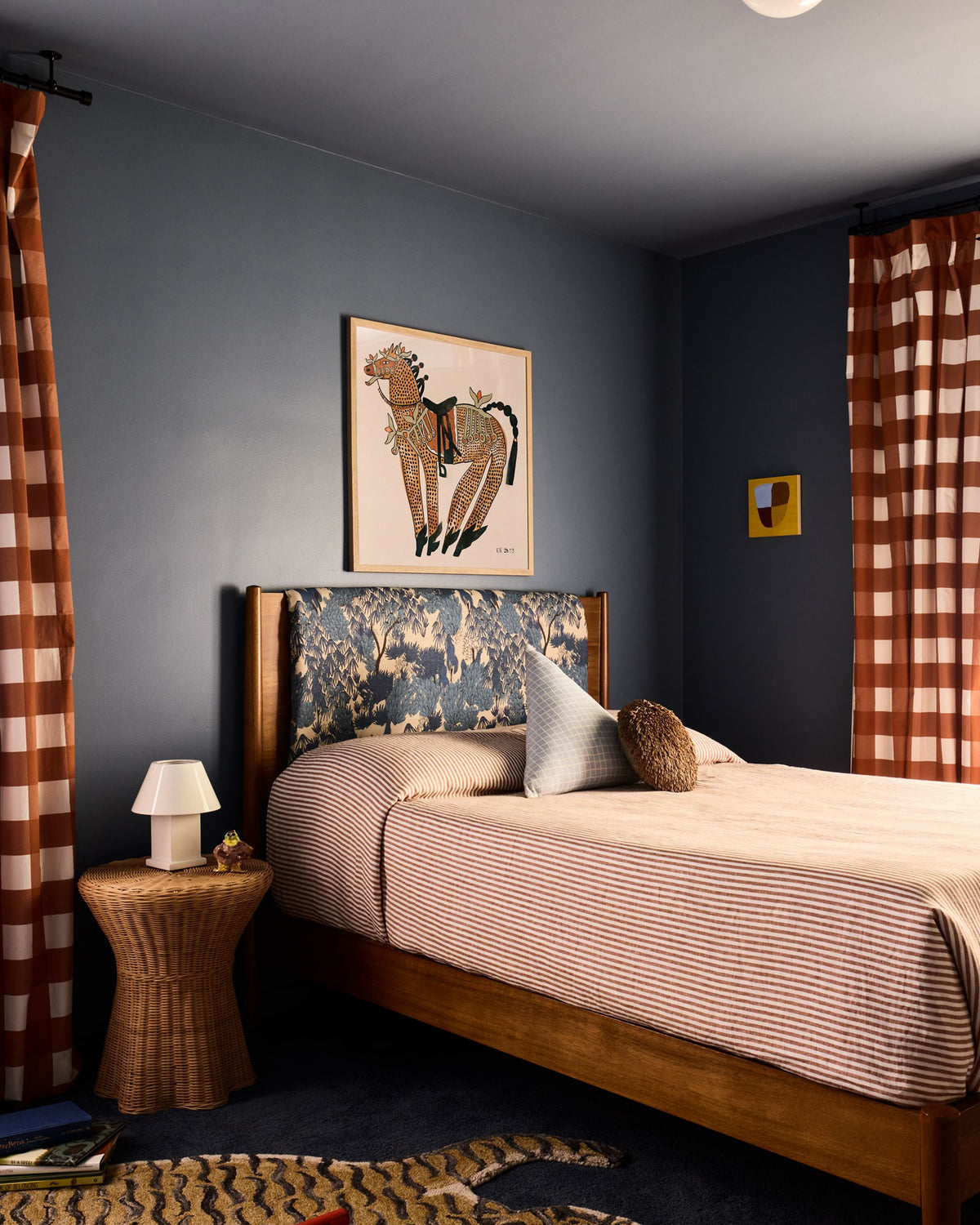 A styled bedroom interior featuring a wooden bed with striped bedding, a patterned upholstered headboard, dark blue walls, checkered curtains, and framed wall art.