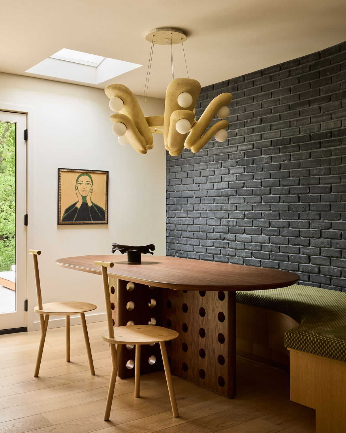 A modern dining area featuring a unique sculptural pendant light in a warm beige wood finish with white spherical accents, hanging above a wooden table and chairs in a room with a dark brick accent wall.