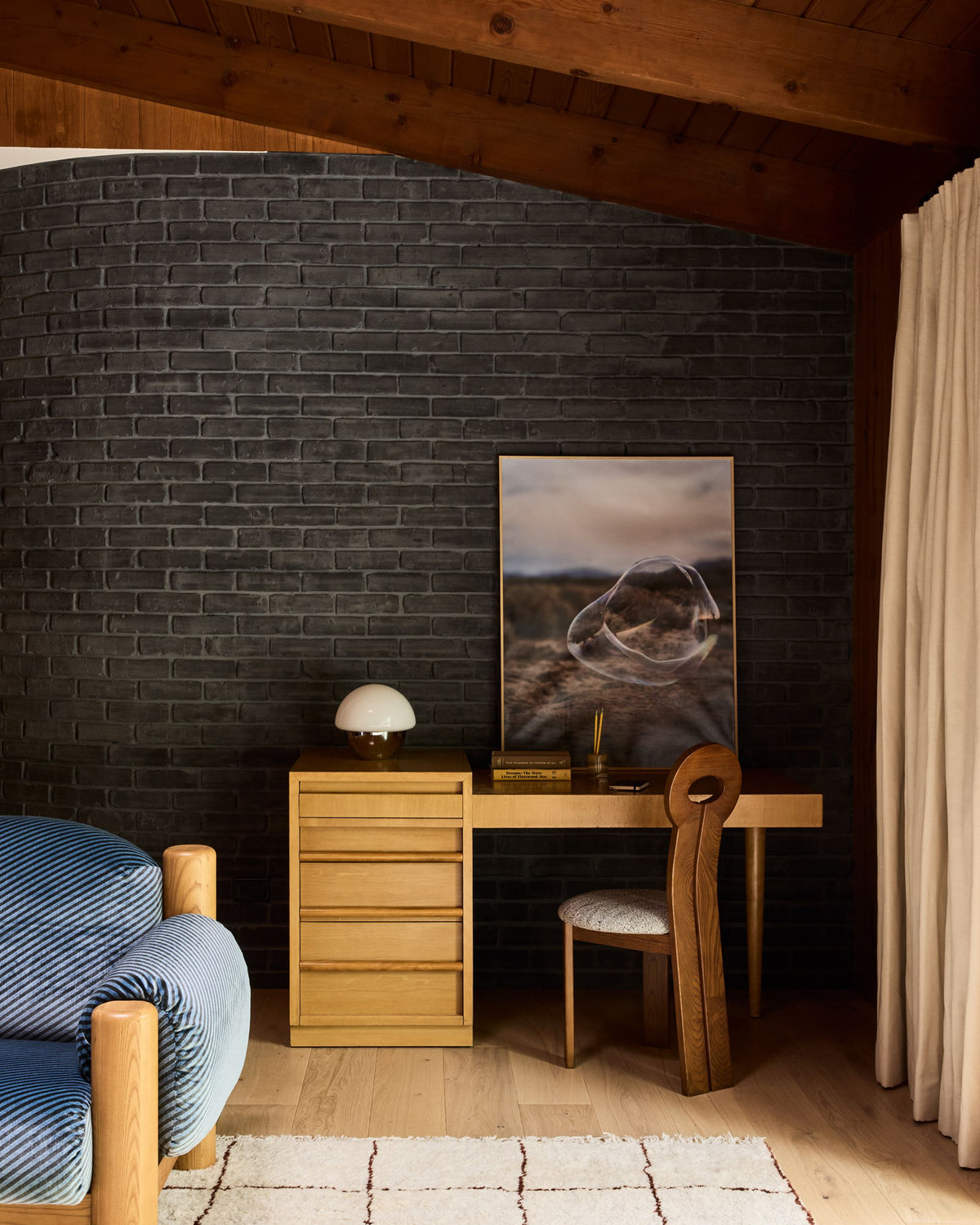 A styled interior corner featuring a dark charcoal brick wall, a light wood desk with a matching chair, a small lamp, and a framed landscape print. A blue upholstered chair is partially visible in the foreground on a white rug, with exposed wooden ceiling beams and a cream curtain at the right.