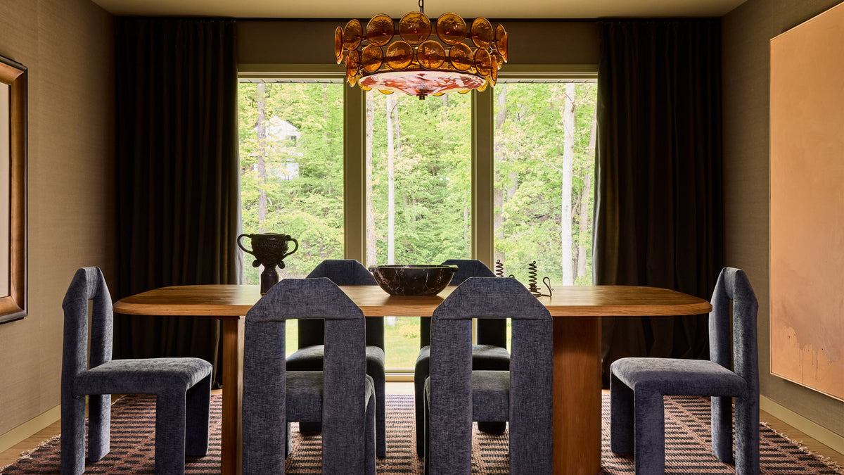 A dining room interior with a wooden dining table, six blue upholstered chairs, a large chandelier, dark curtains, and tall windows looking out onto a forest.