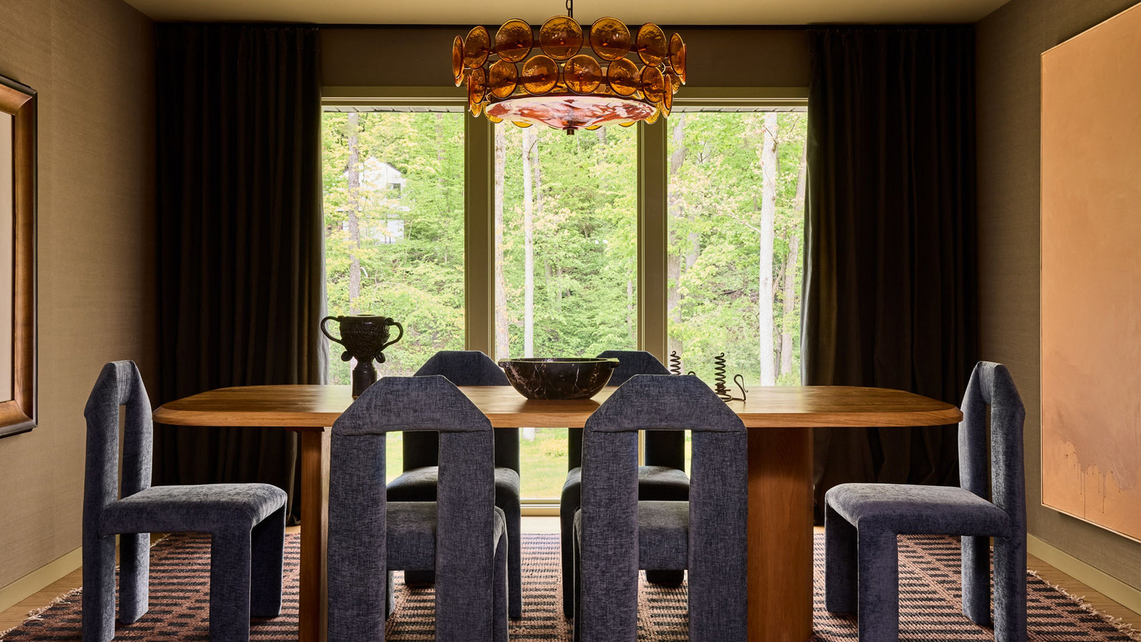 A dining room interior with a wooden dining table, six blue upholstered chairs, a large chandelier, dark curtains, and tall windows looking out onto a forest.