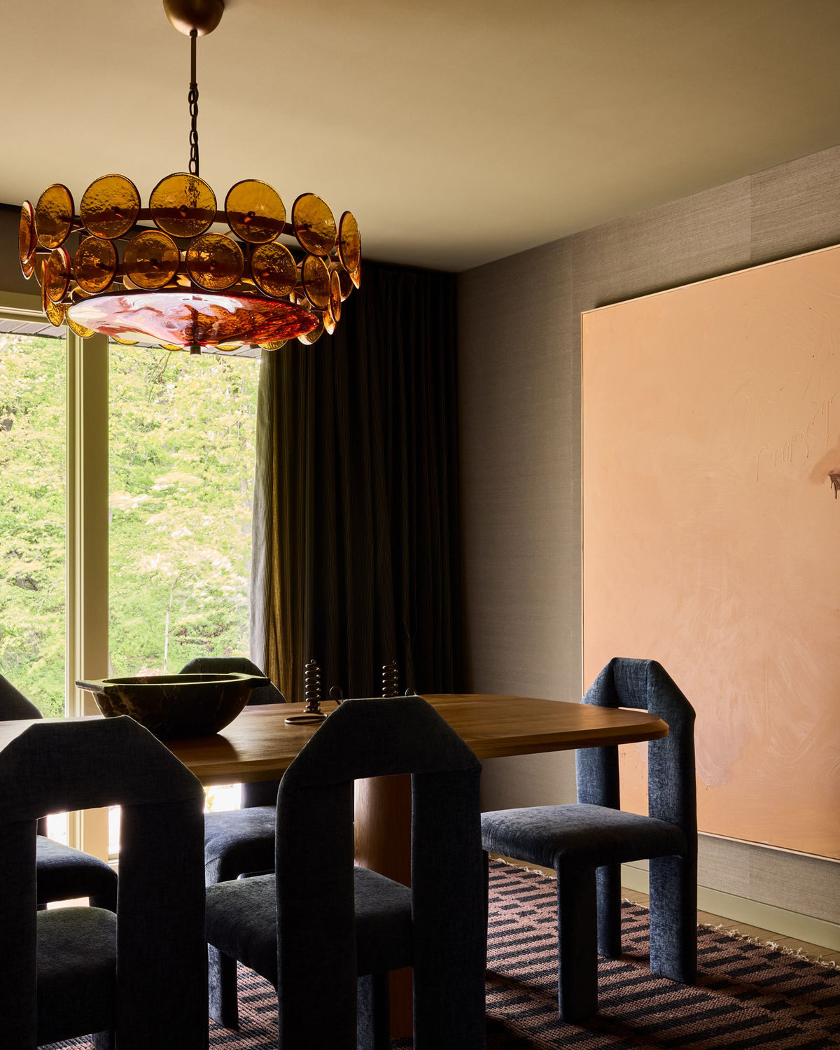 A dining room scene with a large amber glass chandelier hanging above a wooden table and dark upholstered chairs. A tall peach wall art piece and window with greenery outside are visible in the room.