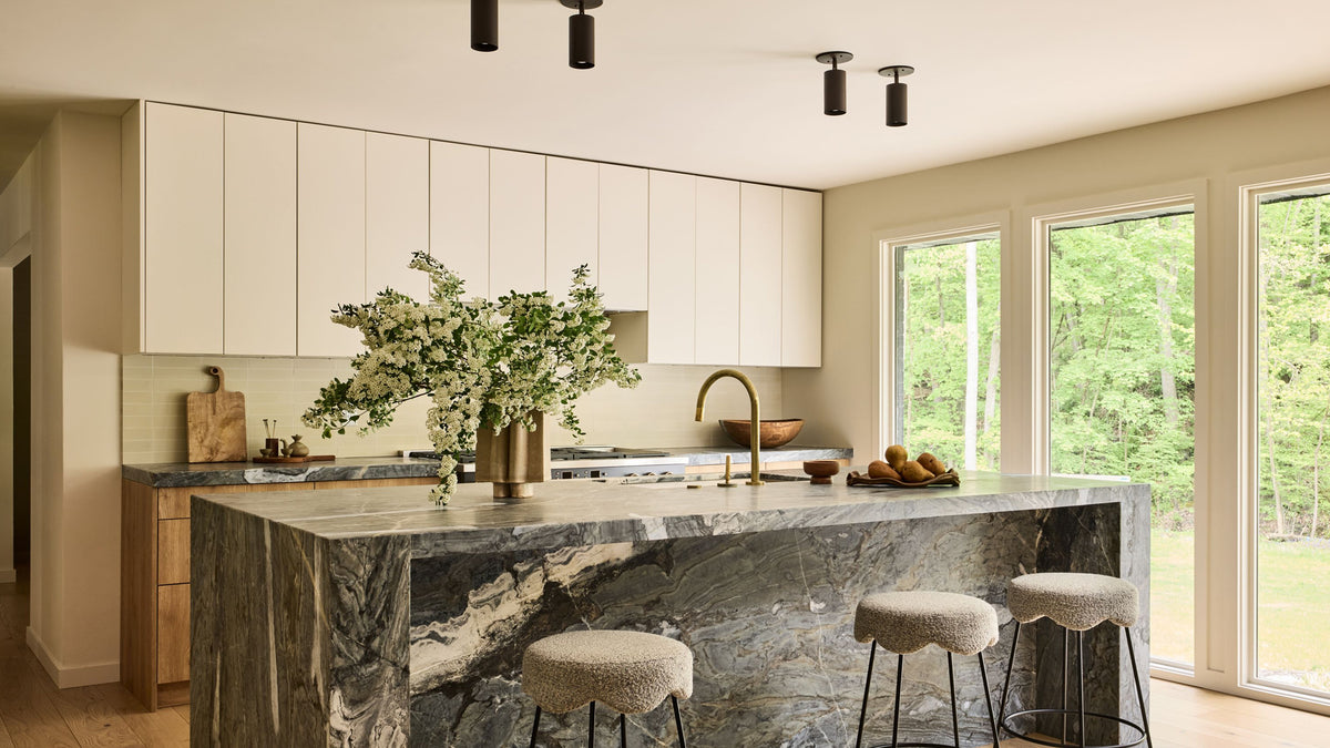 A modern kitchen interior with a large marble island, white cabinets, a gold faucet, and three bar stools. Large windows reveal a green outdoor view.