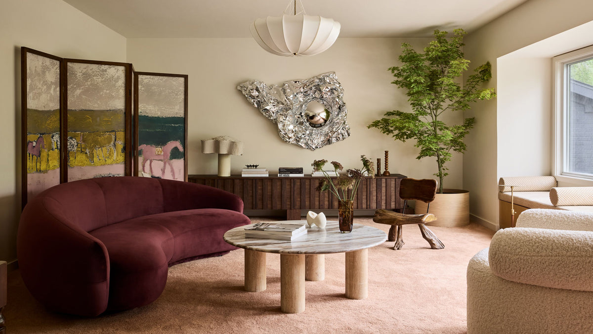 A styled living room interior featuring a curved deep burgundy velvet sofa, a round marble-top coffee table with wooden legs, a beige textured armchair, a potted green tree, and abstract wall art.