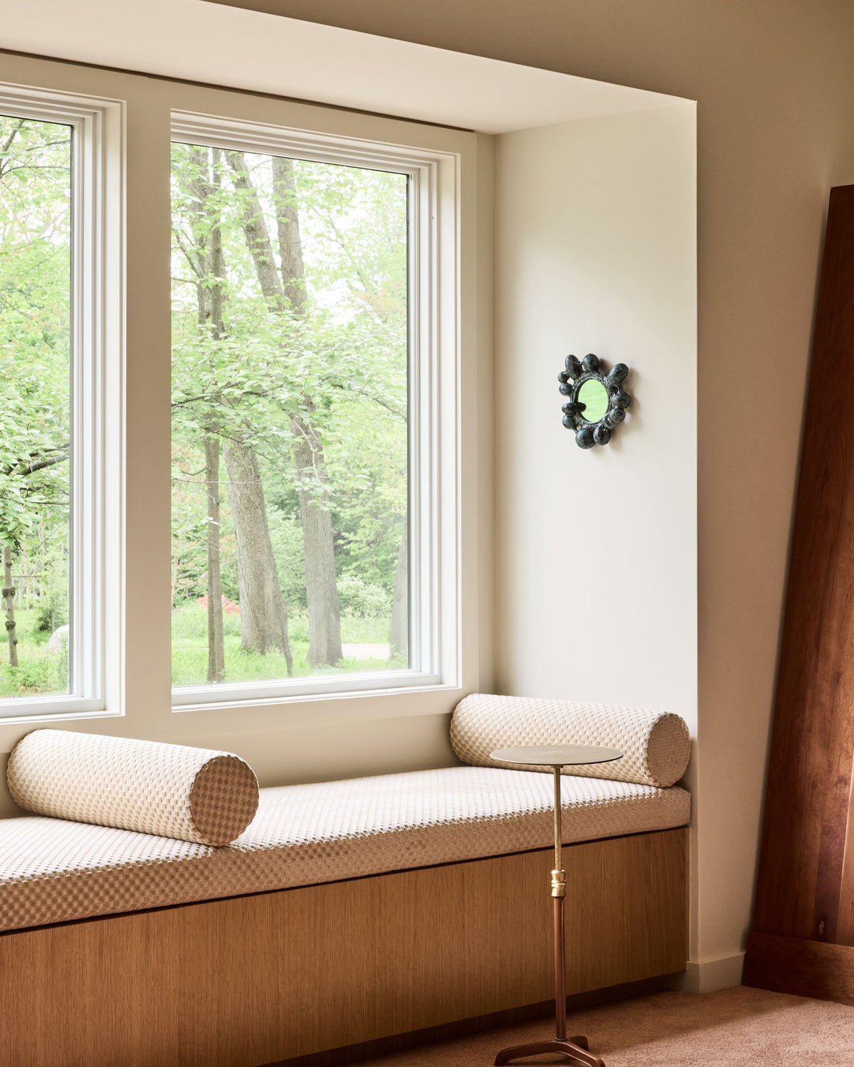 A cozy window seat cushion with cylindrical bolster pillows in a beige and white patterned fabric, set in a warm wood base inside a bright room with a view of green trees outside.