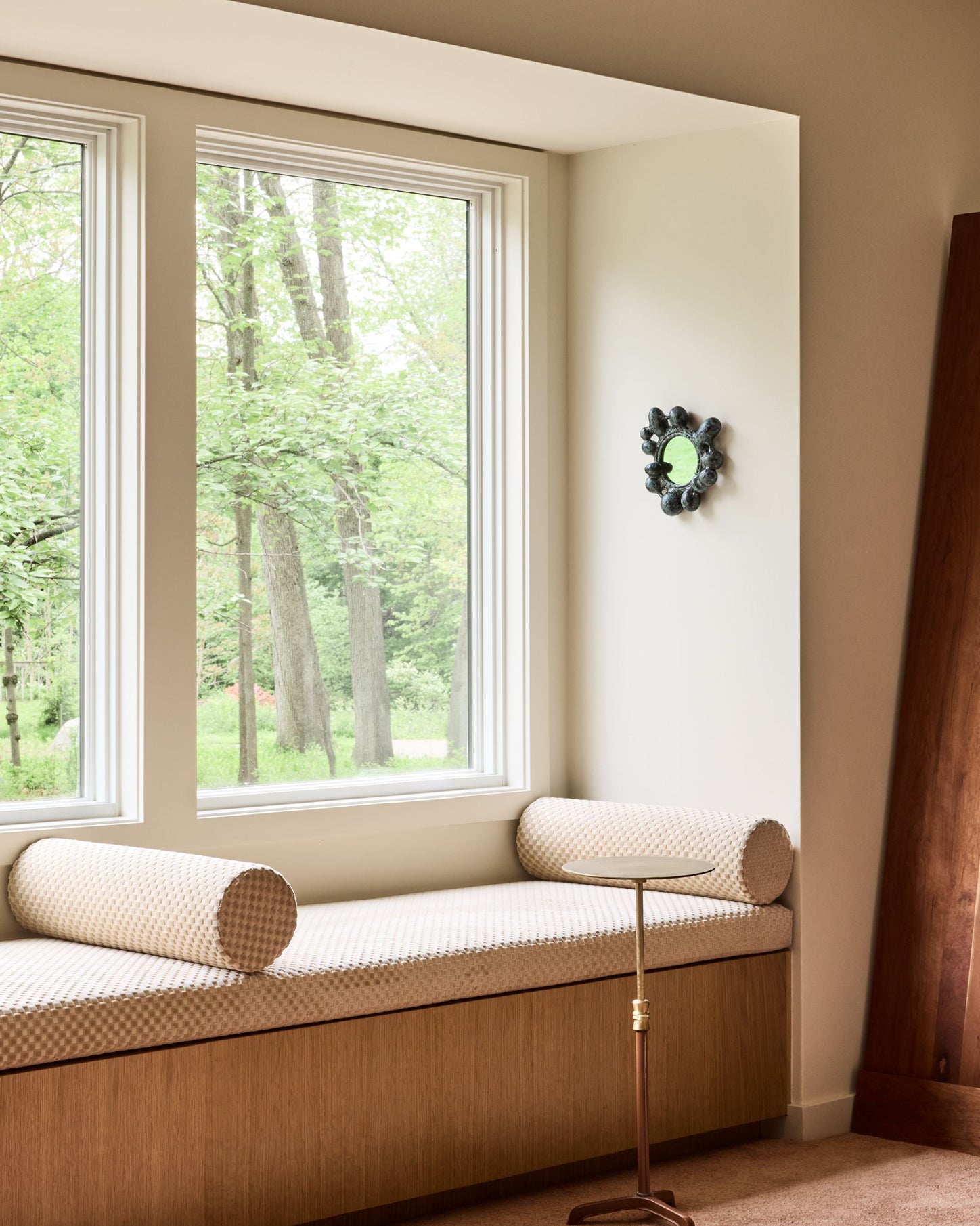 A cozy window seat cushion with cylindrical bolster pillows in a beige and white patterned fabric, set in a warm wood base inside a bright room with a view of green trees outside.