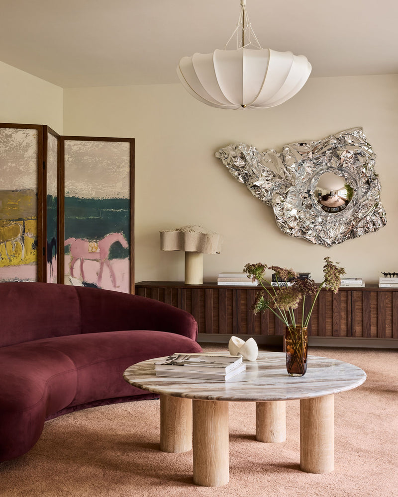 A styled living room interior featuring a curved burgundy velvet sofa, a round marble-top coffee table with wooden legs, a white paper lantern pendant light, and a large organic silver mirror mounted on the wall.