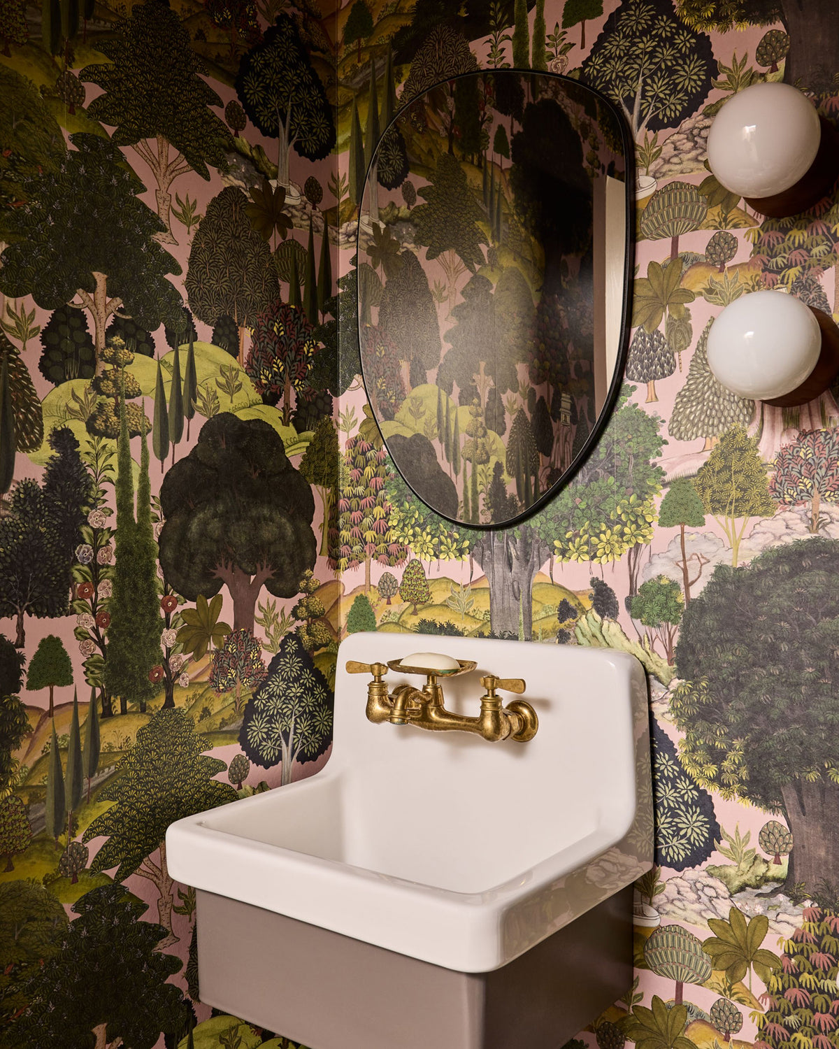 A wall-mounted white ceramic sink with a gold faucet set against a wall covered in dense forest-patterned wallpaper. An oval mirror hangs above the sink, and two round white wall sconces are visible on the right.