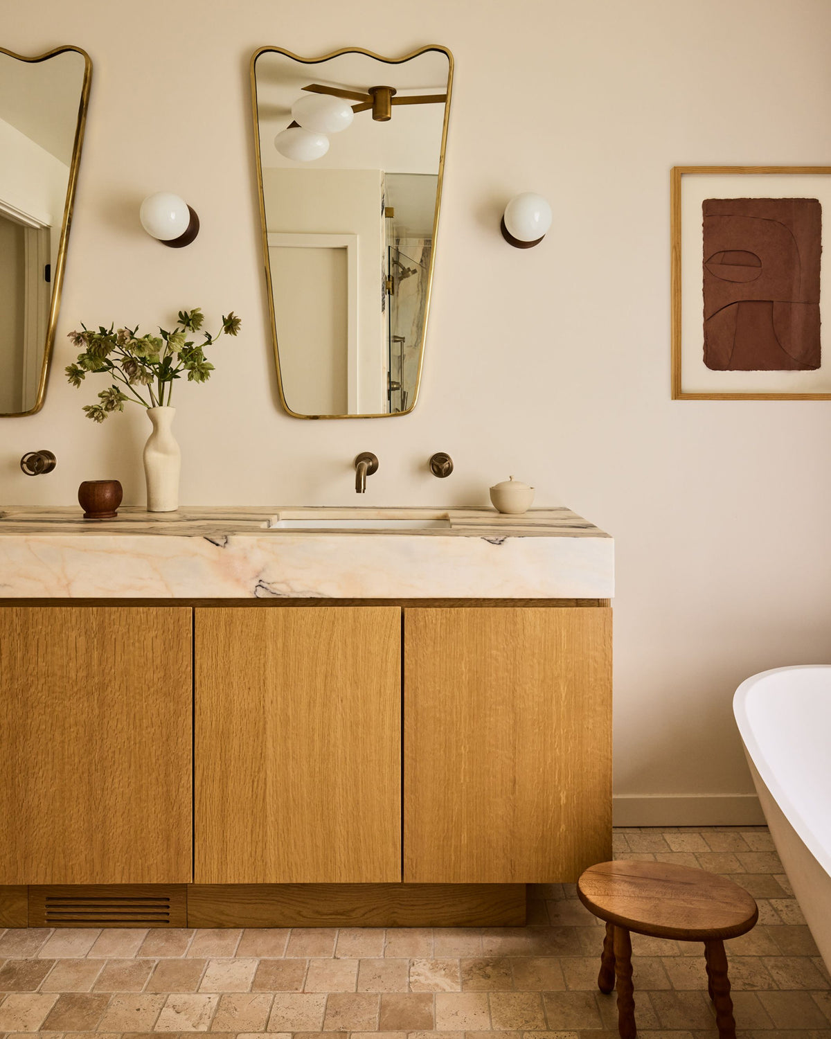 A mid-century modern bathroom vanity featuring light oak wood cabinets, a white marble countertop with an integrated sink, and a wavy gold-framed mirror. The room has beige walls, a small wooden stool, and a white bathtub edge visible on the right.