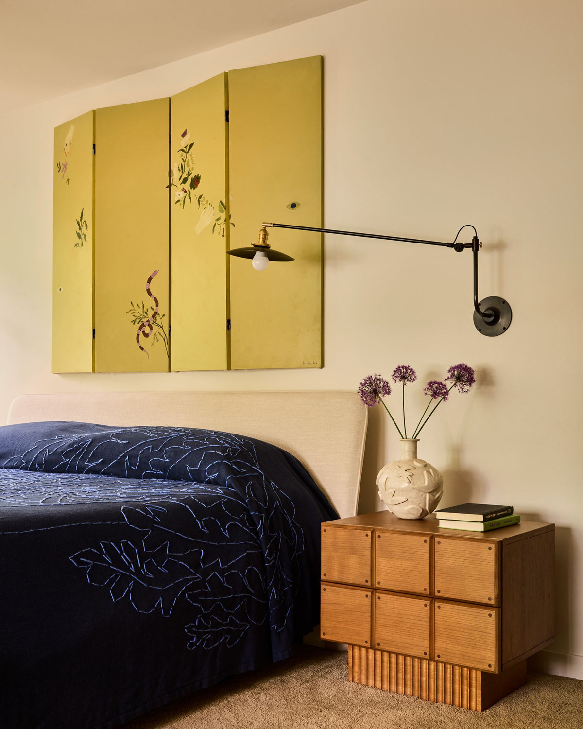 A styled bedroom corner featuring a mustard yellow four-panel folding screen with delicate floral illustrations, a black adjustable wall sconce, a wooden nightstand, and a navy blue patterned bedspread.