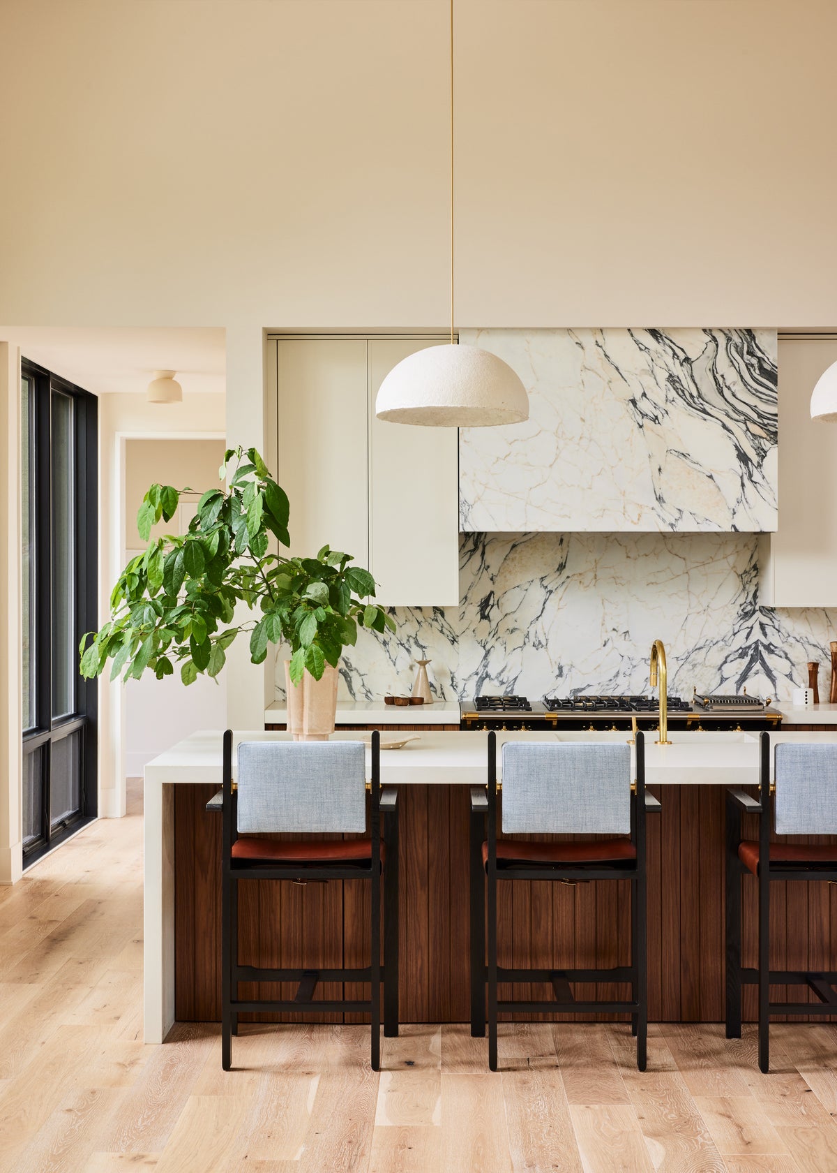 A modern kitchen island with dark wood bar stools featuring light blue upholstered backs, set in a bright kitchen with a marble backsplash, pendant lights, and a potted plant.