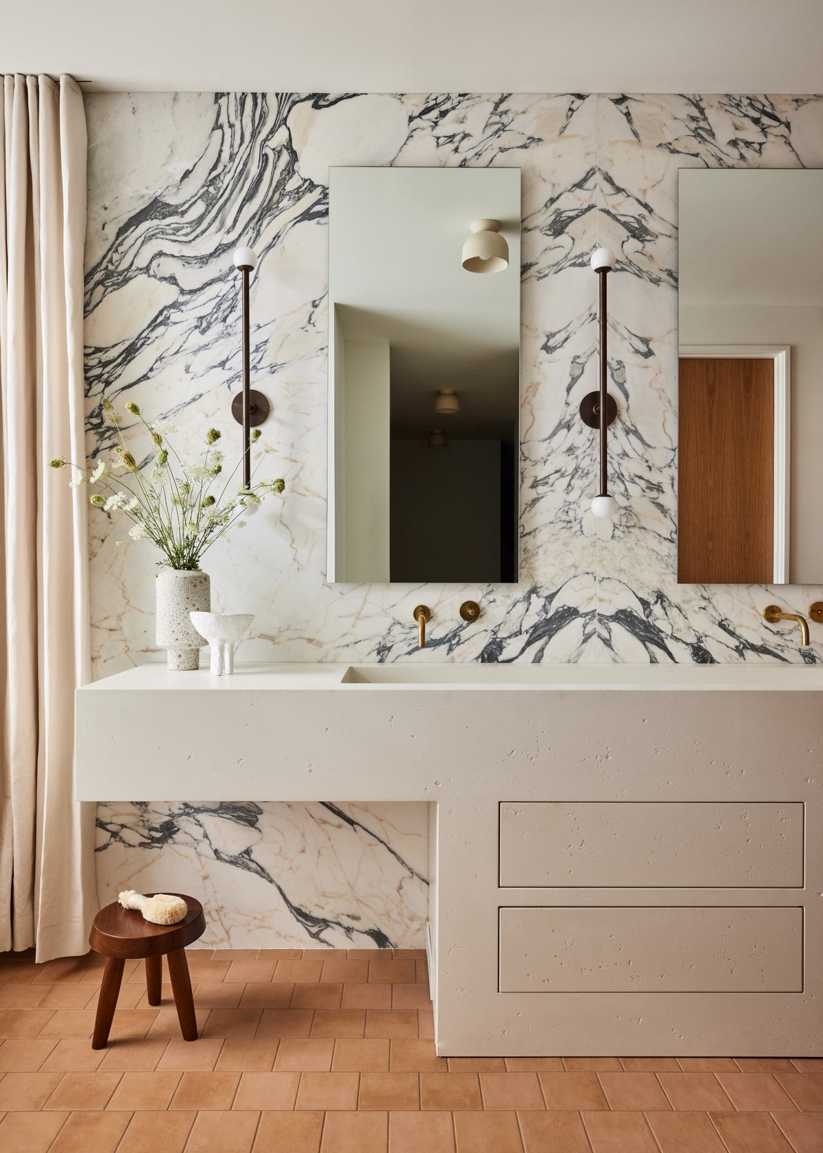 A modern bathroom vanity featuring a white double-sink countertop with a white marble backsplash and wall, two rectangular mirrors, and gold fixtures. A small wooden stool with a vase of flowers sits on the floor next to a beige curtain.