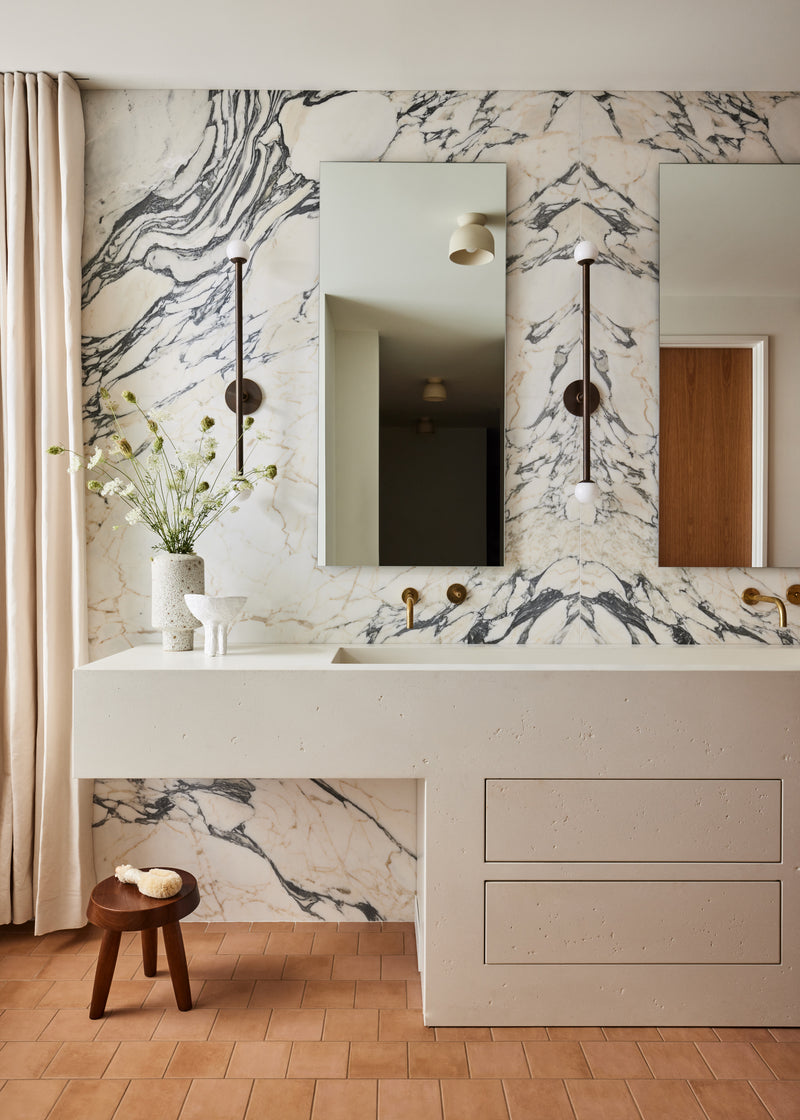 A modern bathroom vanity featuring a white double-sink countertop with a white marble backsplash and wall, two rectangular mirrors, and gold fixtures. A small wooden stool with a vase of flowers sits on the floor next to a beige curtain.