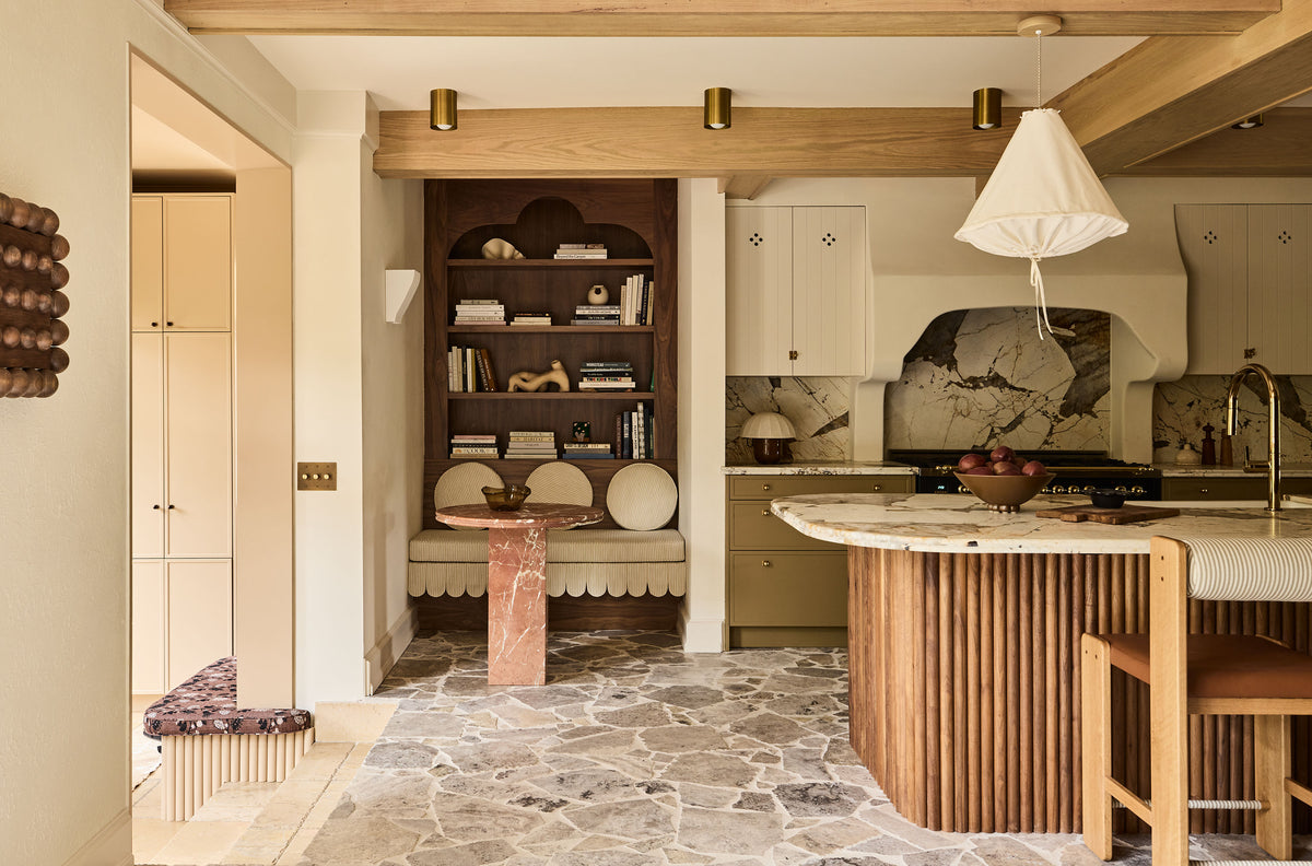 A kitchen interior with light wood cabinetry, a slatted wood island base, a white conical pendant light, stone tile flooring, and a built-in dark wood shelving unit with books and decor.