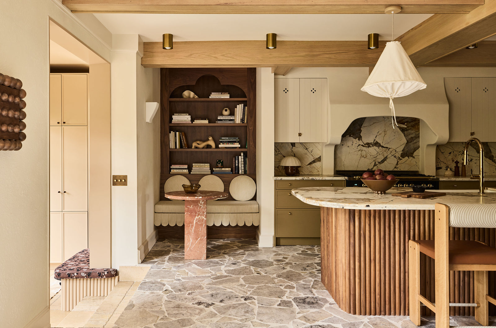 A kitchen interior with light wood cabinetry, a slatted wood island base, a white conical pendant light, stone tile flooring, and a built-in dark wood shelving unit with books and decor.