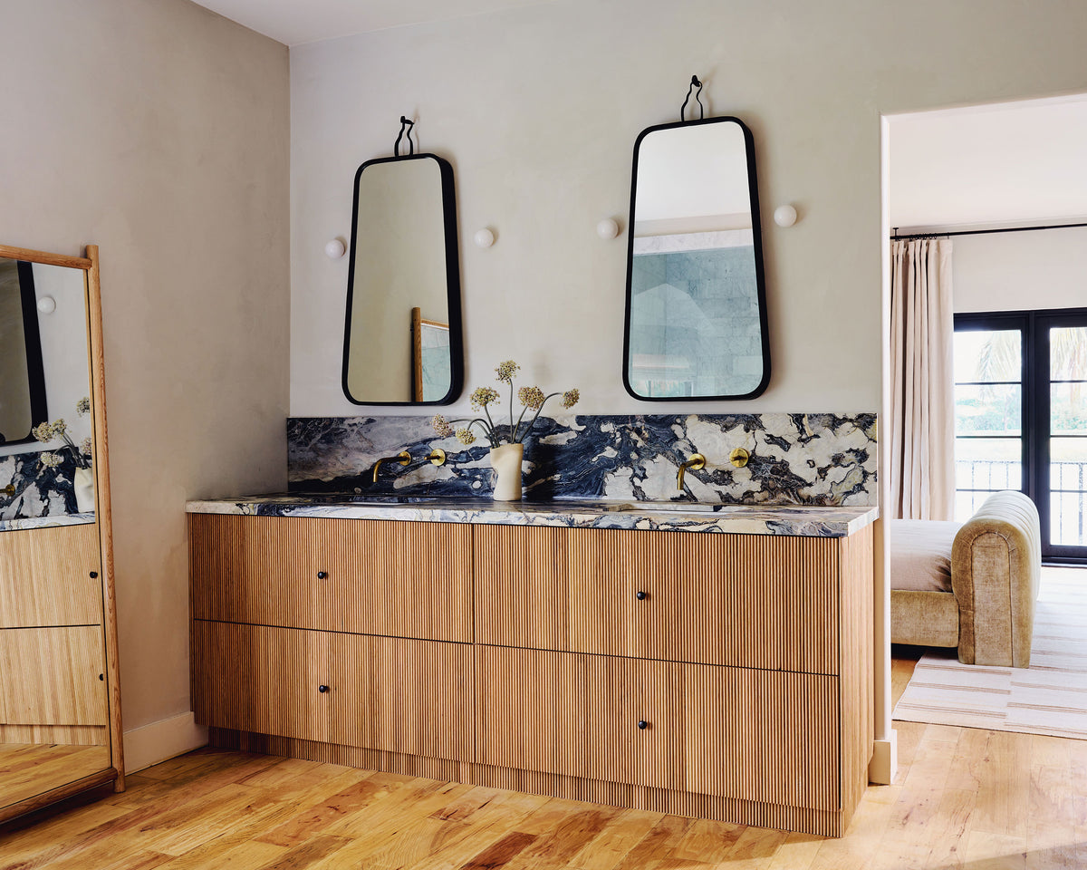 A modern double-sink bathroom vanity with a light wood cabinet, a marble countertop and backsplash, and two black-framed mirrors on a beige wall.