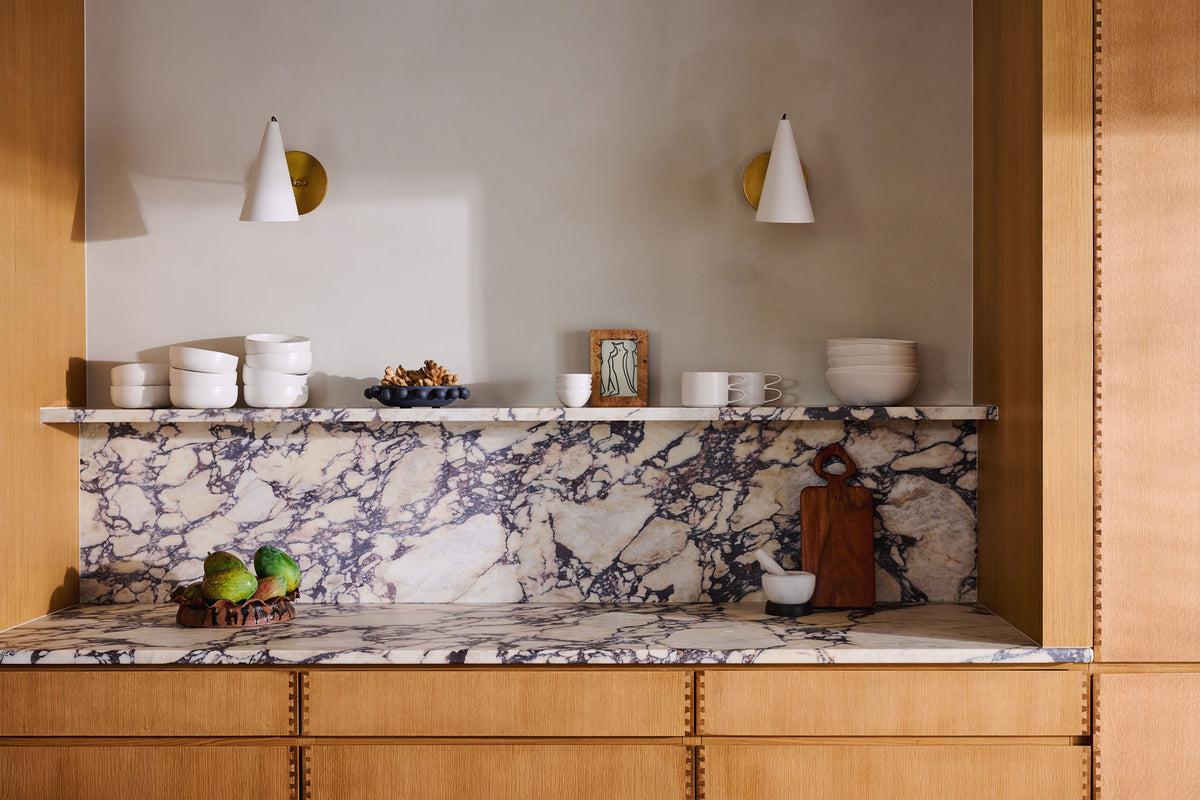 A styled kitchen interior featuring a white marble countertop and backsplash with dramatic dark veining, set in light wood cabinetry. A white shelf above holds ceramic bowls, a small framed picture, and decorative items, under two white cone-shaped wall sconces.