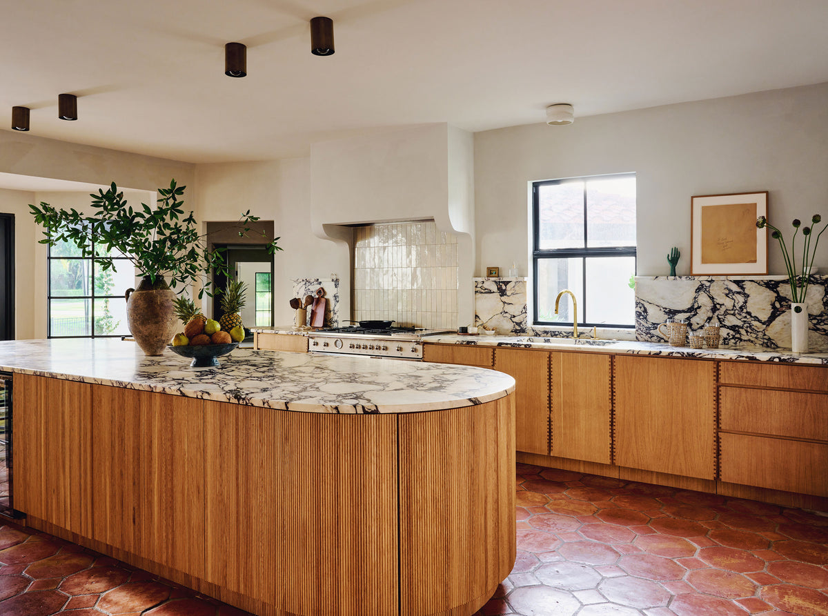A modern kitchen interior with a curved island featuring light wood grain fronts and a white marble countertop, set against cream walls and terracotta tile flooring.