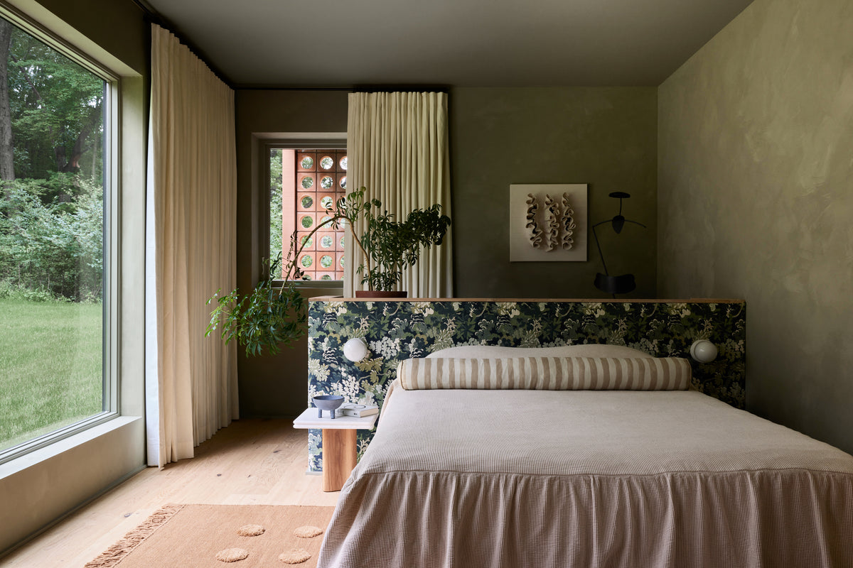 A styled bedroom interior featuring a bed with a floral headboard, beige bedding, a striped pillow, and a large window looking out onto green grass and trees.
