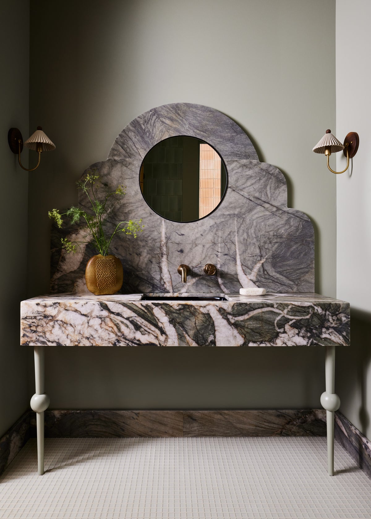 A bathroom vanity featuring a sculptural cloud-shaped marble mirror and a matching marble console table with legs, set against a muted sage green wall with wall sconces.