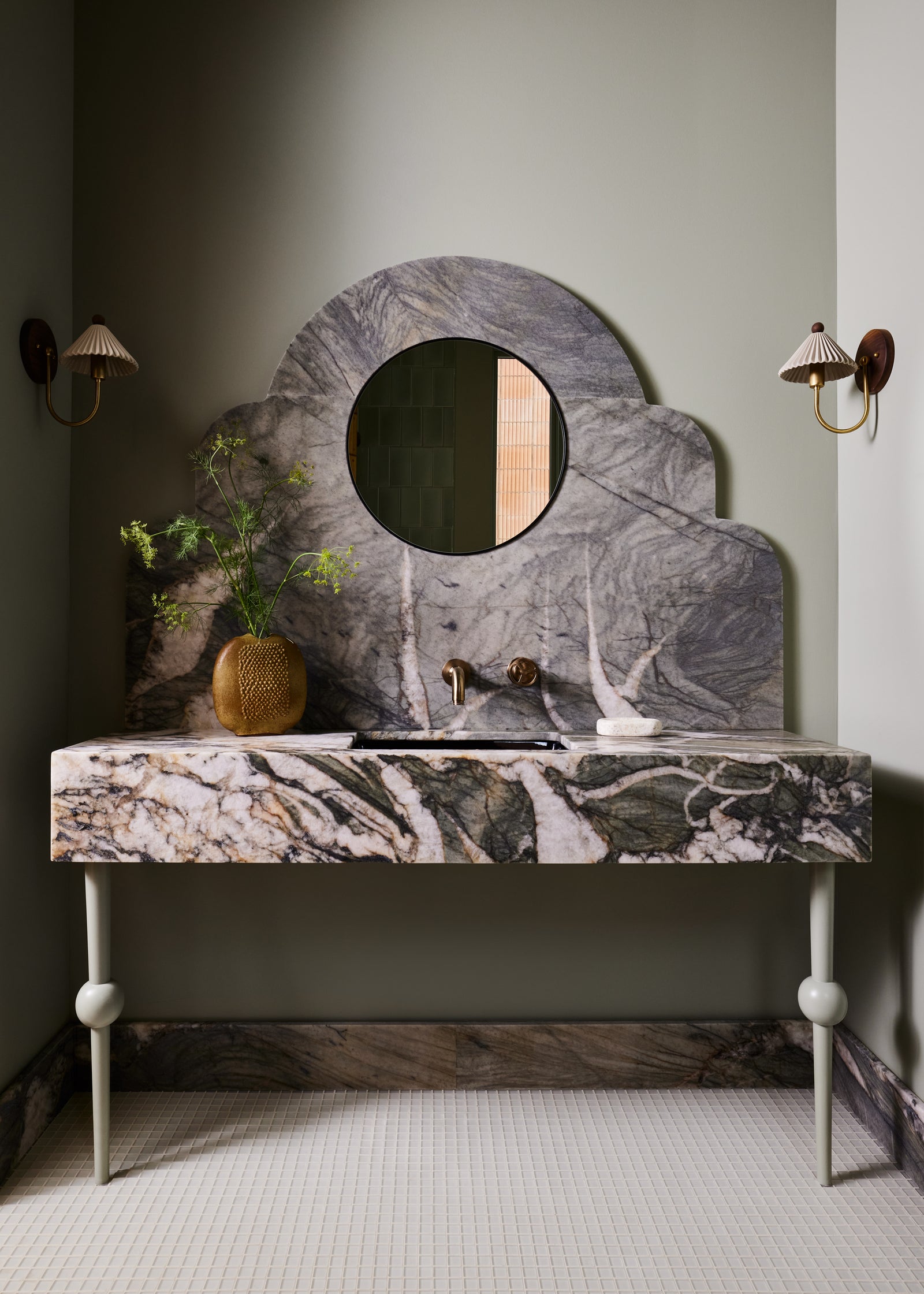 A bathroom vanity featuring a sculptural cloud-shaped marble mirror and a matching marble console table with legs, set against a muted sage green wall with wall sconces.