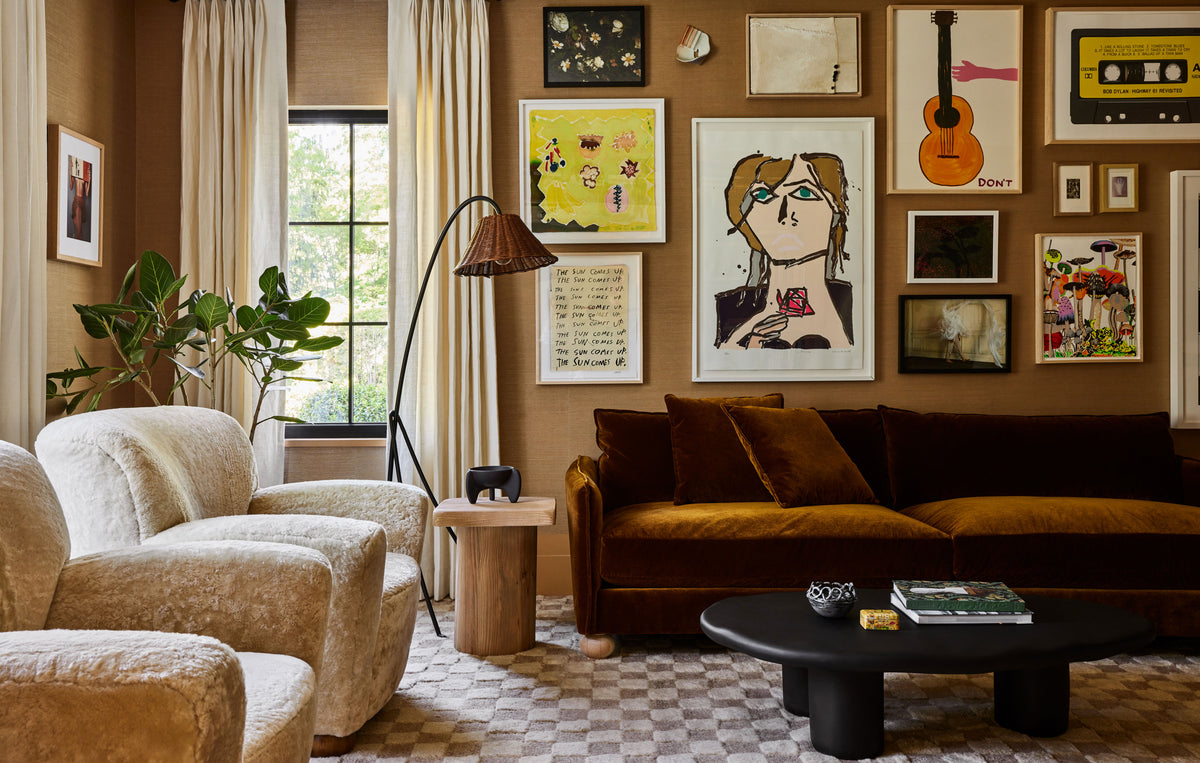 A styled living room interior with a brown velvet sofa, cream boucle armchairs, a black coffee table, and a warm brown wall covered in framed art, including a guitar print and cassette tape artwork.
