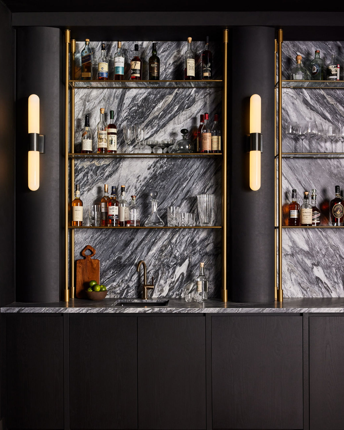 A luxury bar setup featuring a black marble backsplash with gold-framed shelves stocked with bottles and glassware, flanked by two black columns with vertical wall sconces.