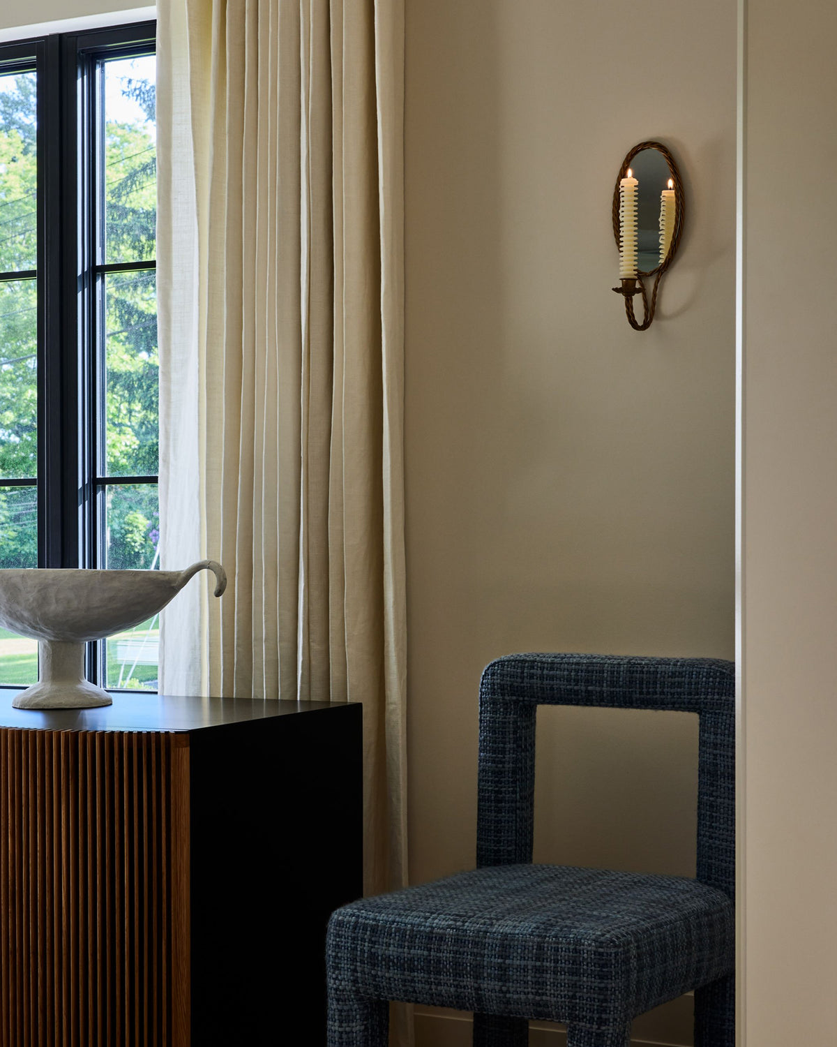 A wall sconce with two lit candle-style bulbs mounted on an ivory wall in a styled interior corner with a blue upholstered chair, cream curtains, and a window.