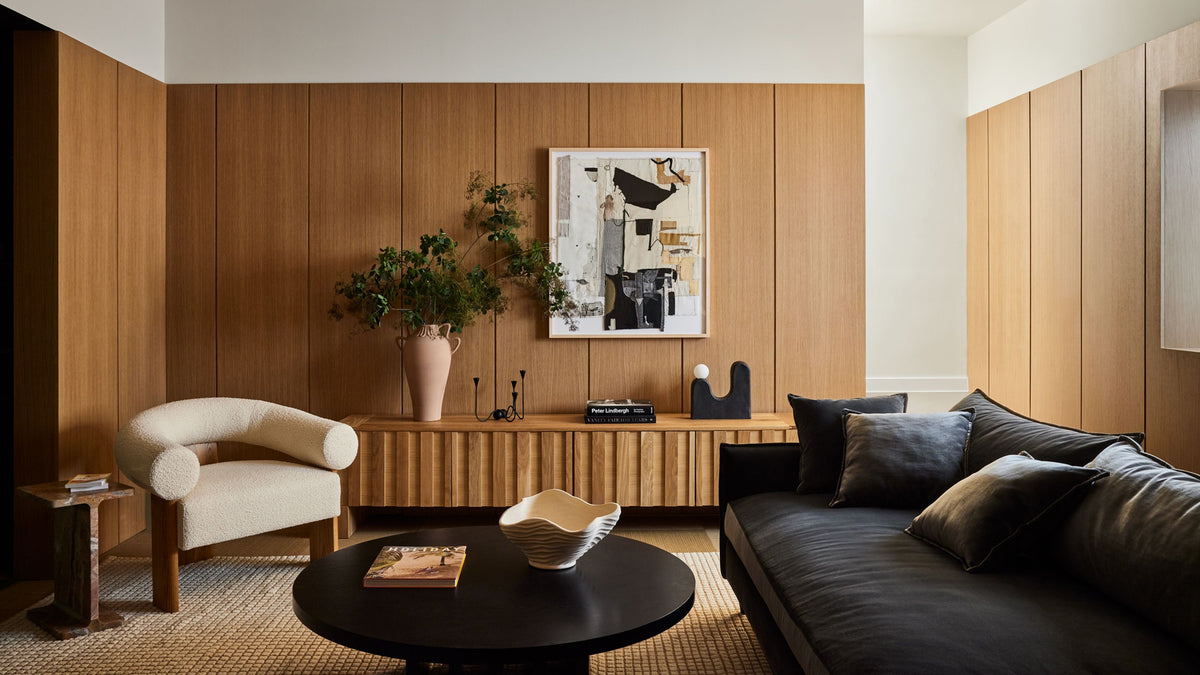 A modern living room interior with warm wood-paneled walls, a cream curved armchair, a dark charcoal sofa with pillows, a round black coffee table, and framed wall art above a Fort Credenza by Sarah Sherman Samuel.