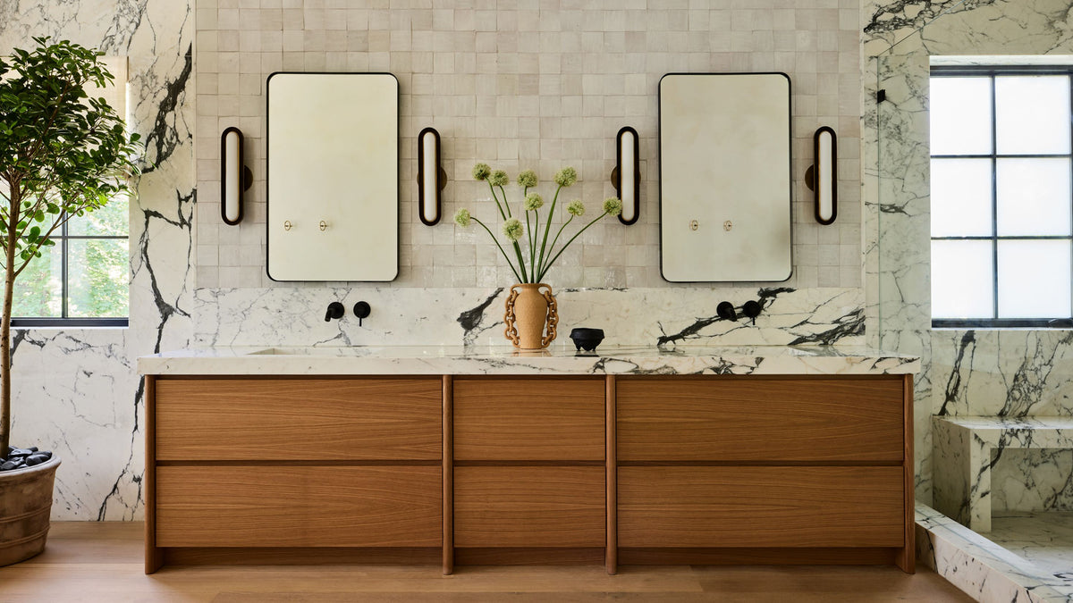 A modern bathroom vanity featuring a light oak wood vanity, a white marble countertop with a central vase of green stems, and two large rectangular mirrors mounted on a tiled wall.