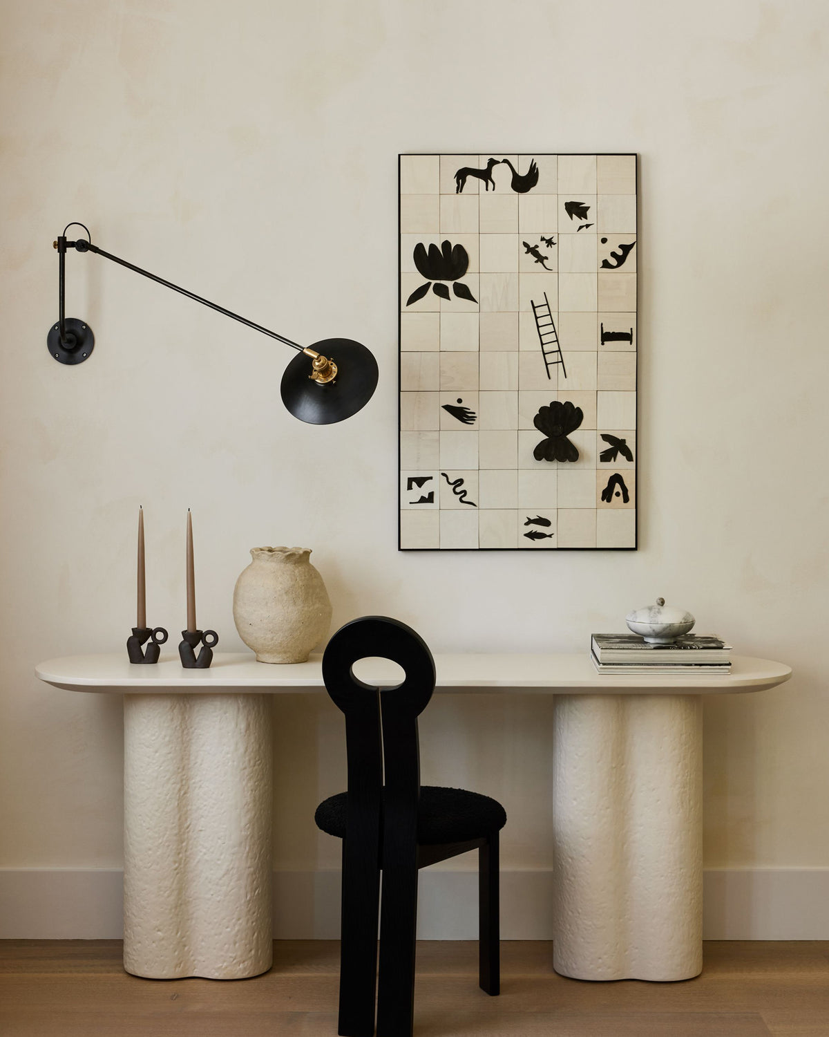 A styled interior scene with a cream console table, textured cylindrical legs, a black chair, a black wall lamp, and a framed artwork by artist Sarah Matthews on a warm ivory wall.