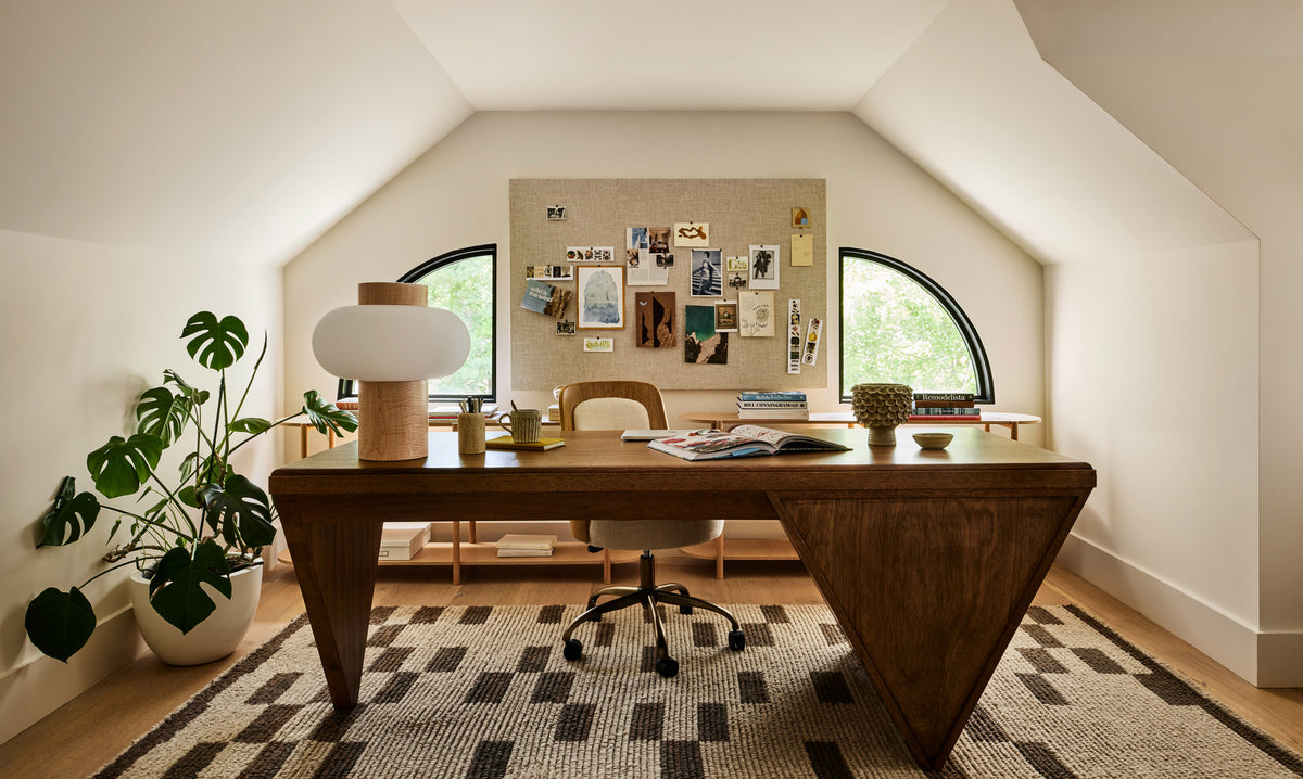 A modern wooden desk with a triangular base sits in a bright, sloped attic home office with arched windows, a corkboard wall, and a patterned rug.