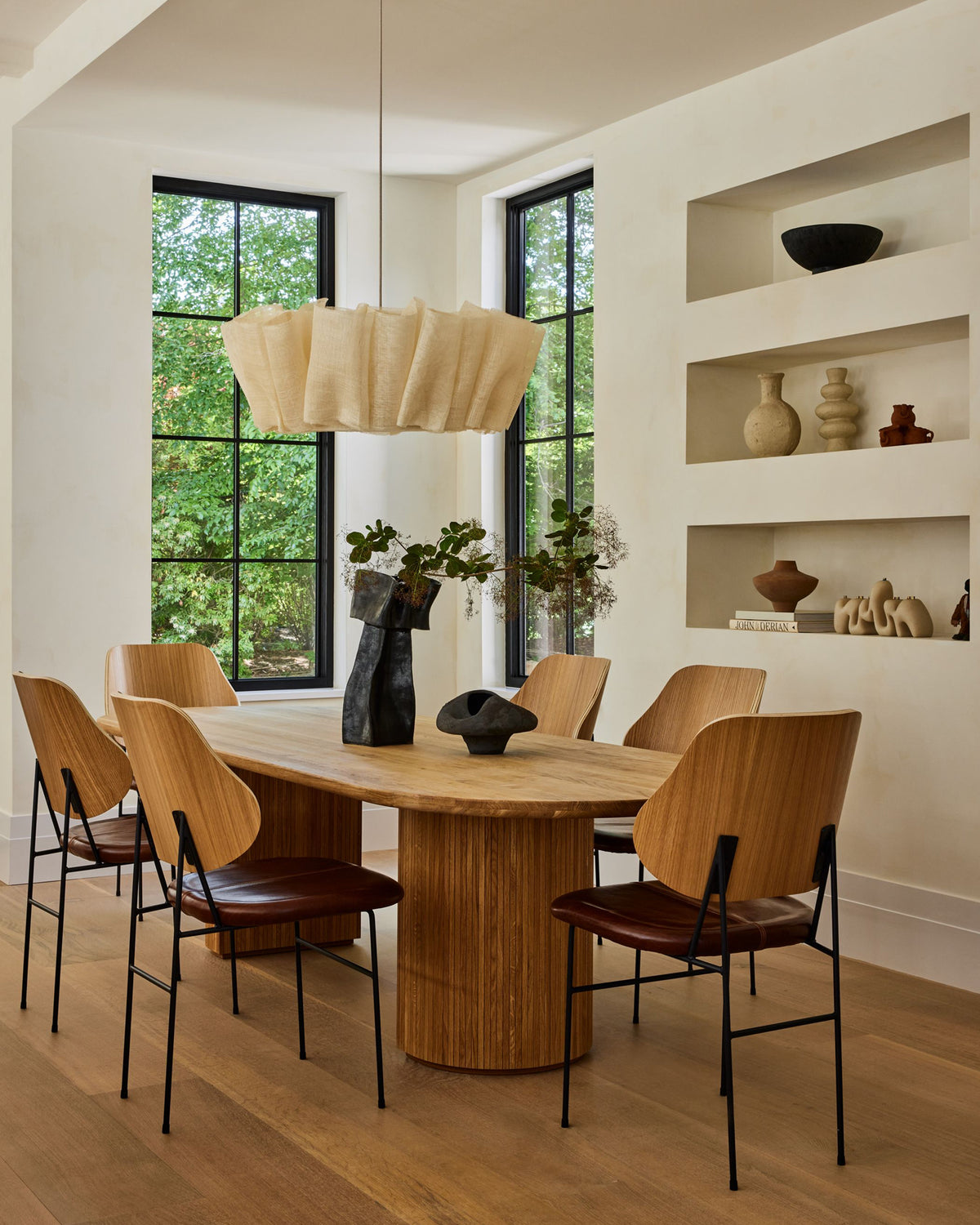 A modern dining room featuring a light wood oval table with a fluted pedestal base, surrounded by six chairs with light wood backs, dark brown leather seats, and black metal frames. A large, pleated beige pendant light hangs above.