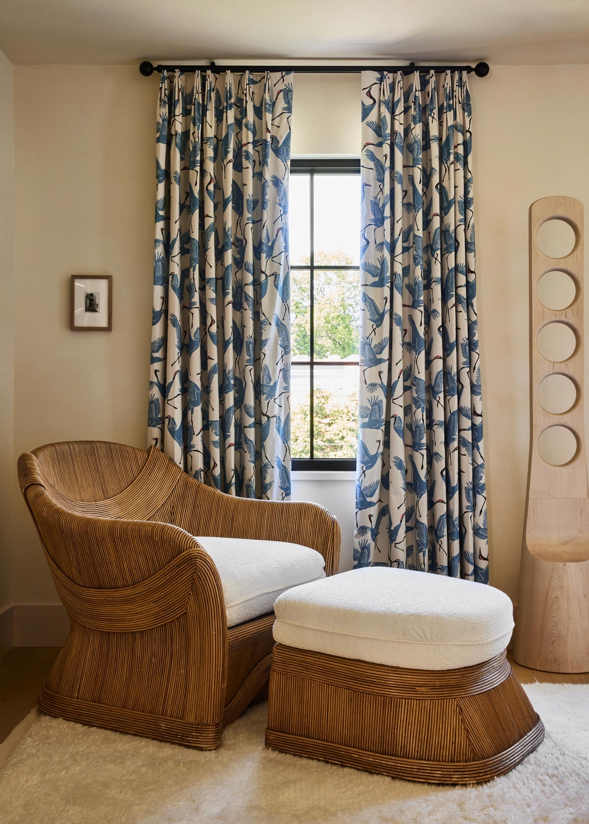 A styled interior room featuring a curved wooden armchair with a white cushion and a matching wooden ottoman with a white cushion, positioned in front of patterned blue and beige curtains hanging over a window.