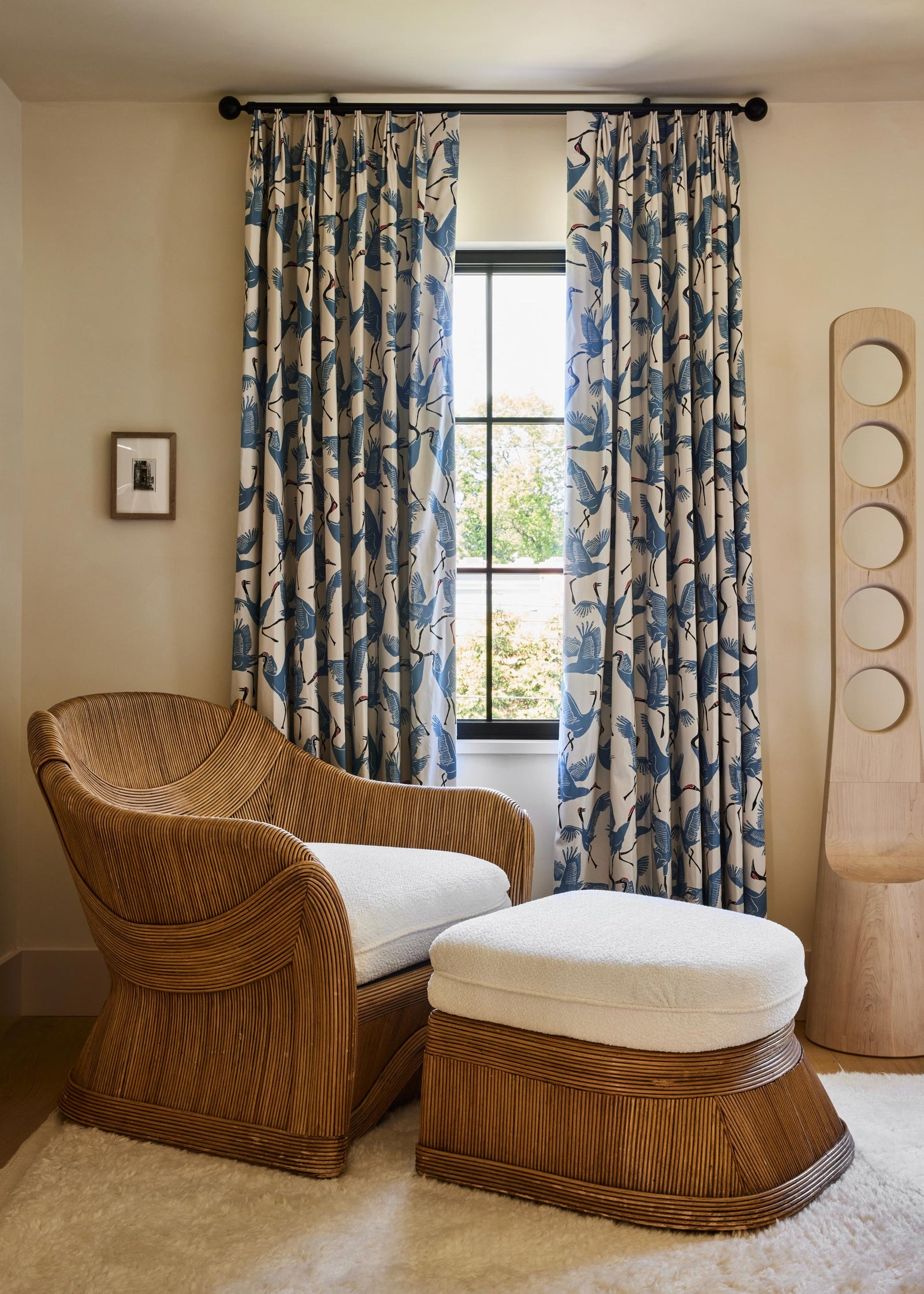 A styled interior room featuring a curved wooden armchair with a white cushion and a matching wooden ottoman with a white cushion, positioned in front of patterned blue and beige curtains hanging over a window.