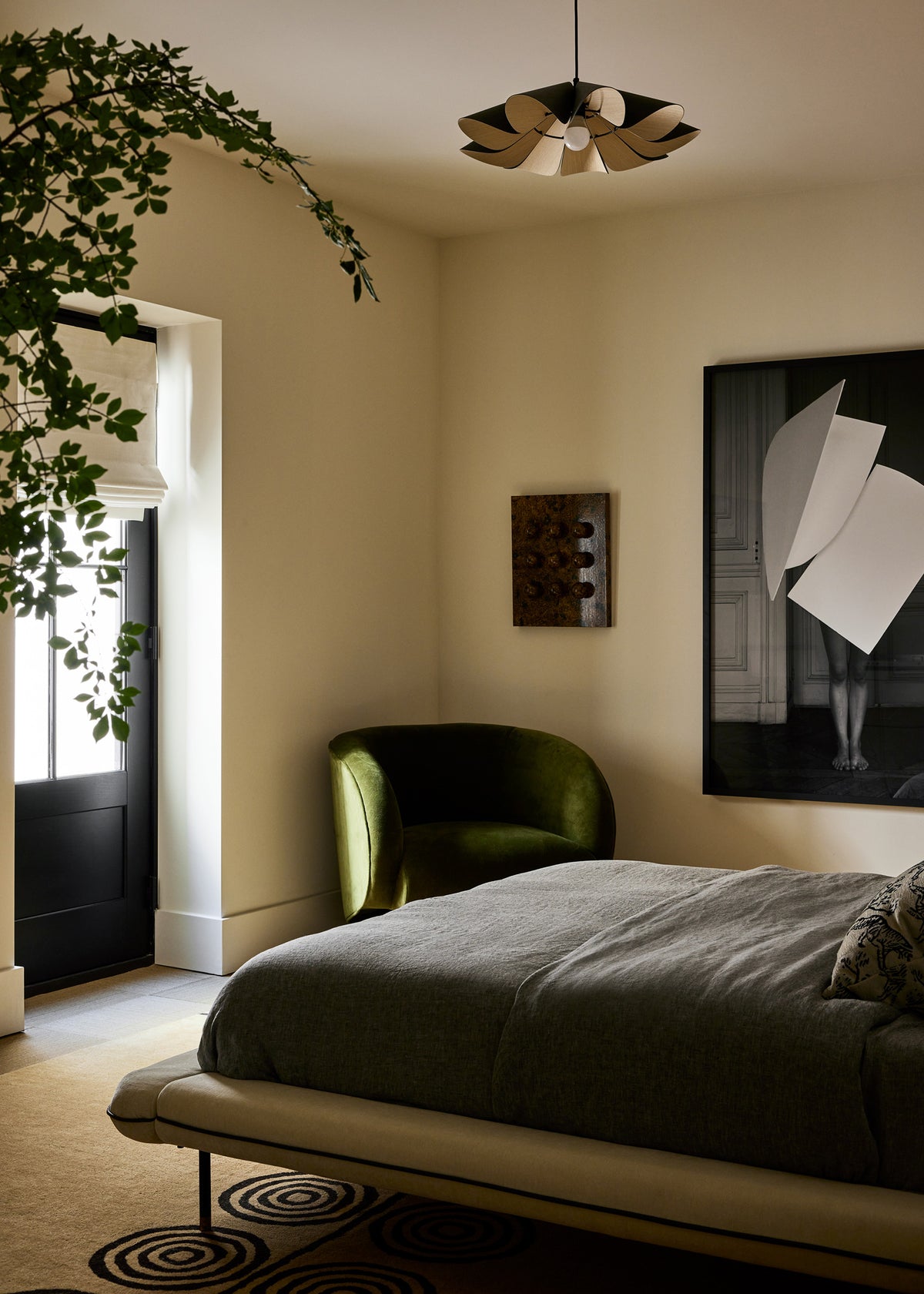 A bedroom interior with a low bed dressed in dark gray bedding, a green velvet accent chair, a black-framed abstract wall art print, and a hanging pendant light in a warm neutral room.