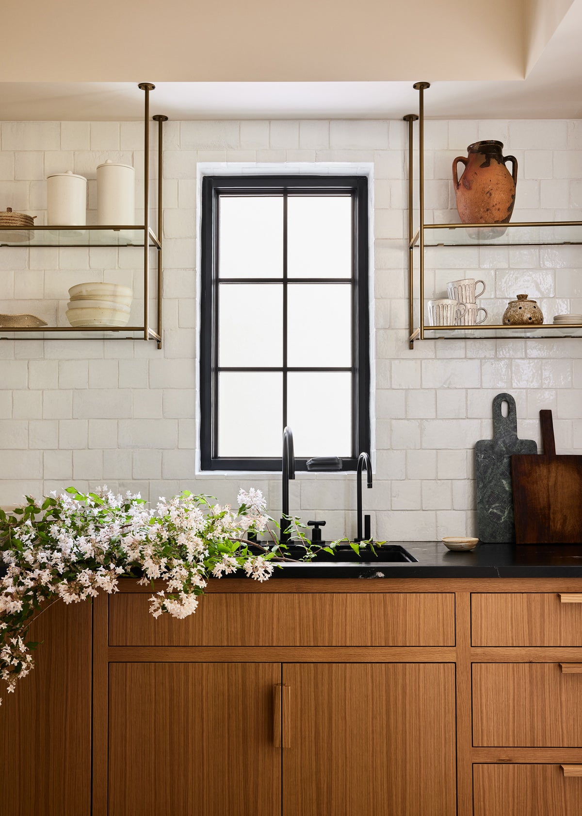 A modern butlers pantry scene with a black-framed window above a black countertop and sink, set in warm wood cabinets. White subway tiles line the wall, with brass floating shelves holding ceramic vessels and a terracotta pot.
