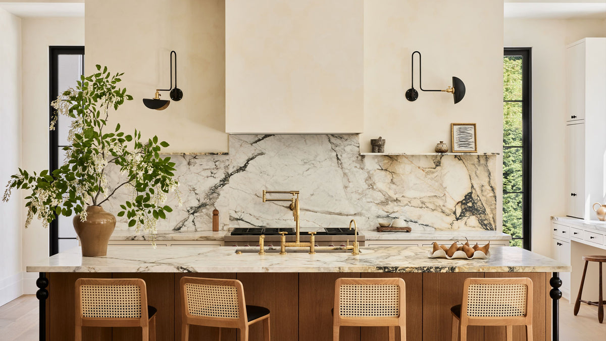 A modern kitchen island with a white marble countertop and backsplash, featuring a brass faucet and sink, wooden cabinetry, four woven-back bar stools, and a large potted green plant on the left.