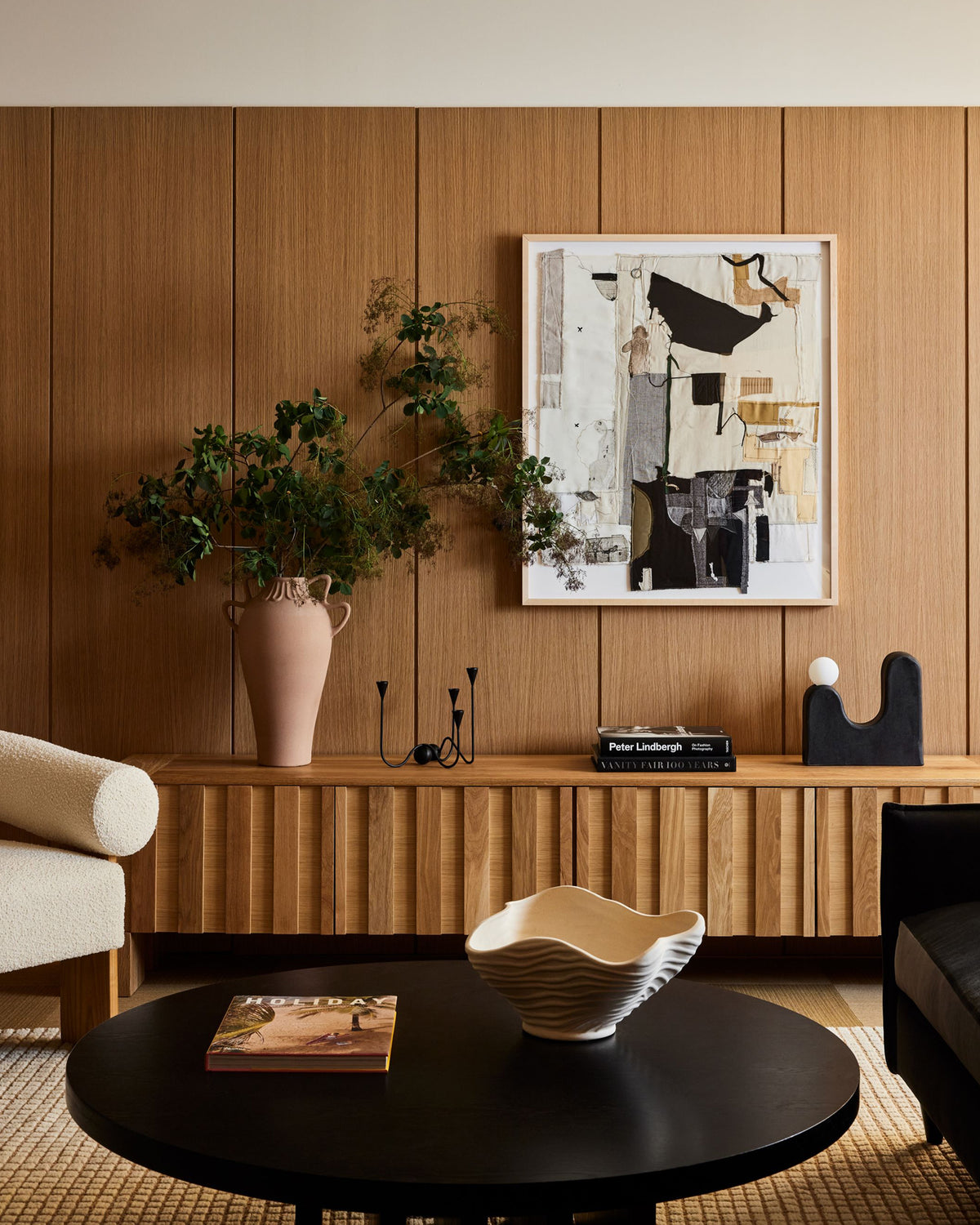 A styled interior scene with a wood-paneled wall, framed abstract artwork, a low wooden console, a large vase with greenery, a black coffee table, and partial seating in a living room setting.