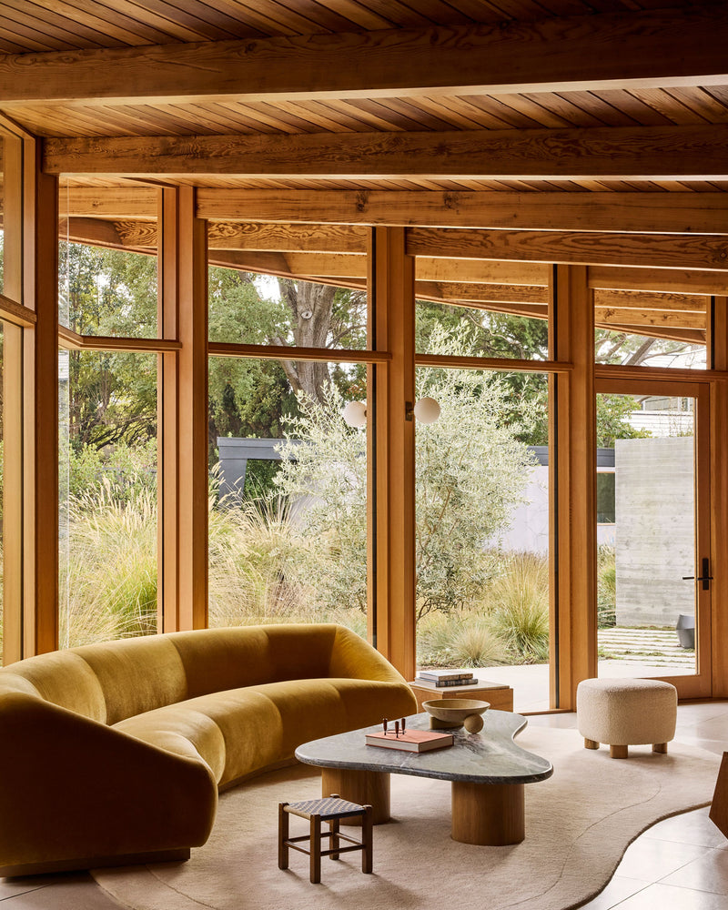 A living room interior with a curved mustard-yellow velvet sofa, a stone-top coffee table, a small stool, and large wood-framed windows overlooking a garden. The room is dominated by warm wood tones and natural light.