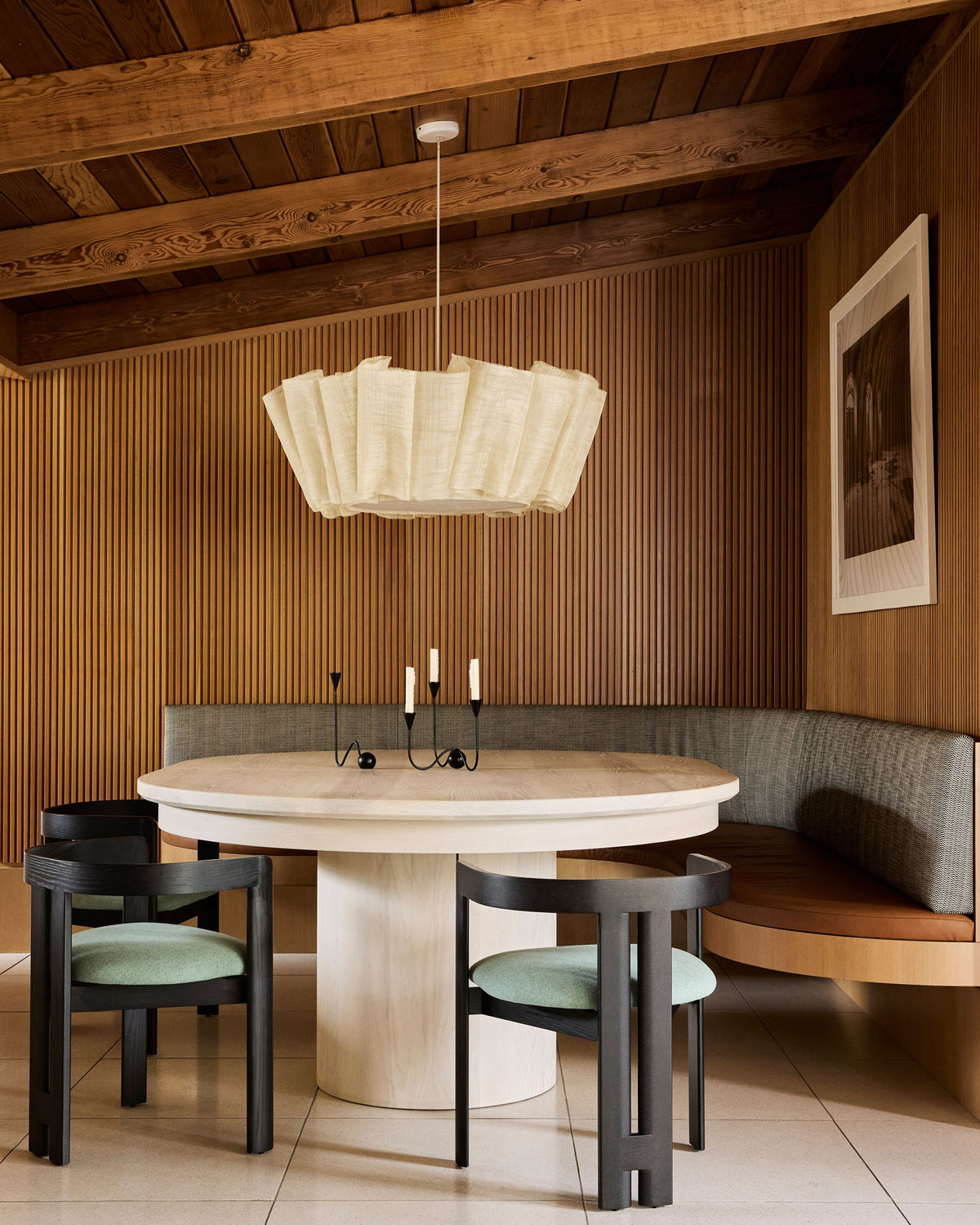 A modern dining room scene featuring a round light wood table, four black chairs with mint green cushions, a curved gray upholstered wood banquette, a large white pleated pendant light, and a warm wood slatted wall and ceiling.